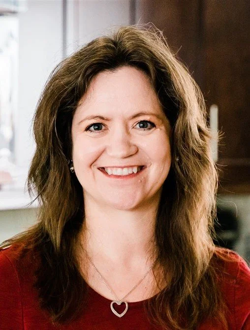 Smiling woman with long brown hair wearing a red top and heart-shaped necklace in an indoor setting.