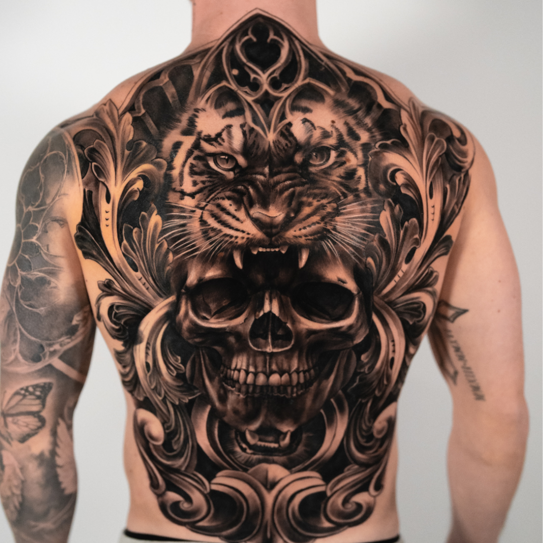 A person's back tattoo featuring a fierce tiger's face above a skull, with ornate decorative elements.