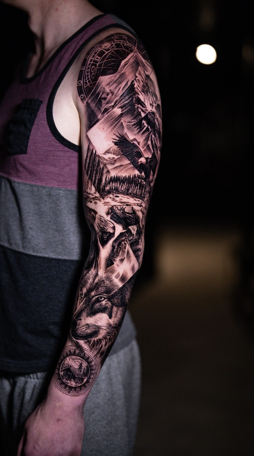 Person with detailed black and grey nature-themed sleeve tattoo featuring mountains, trees, a wolf, an eagle, and compass elements.