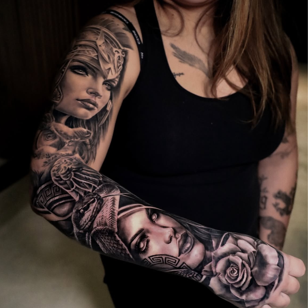 Close-up of a woman's arm with detailed black and gray tattoo sleeve featuring portraits of women, a snake, a rose, and various artistic designs.