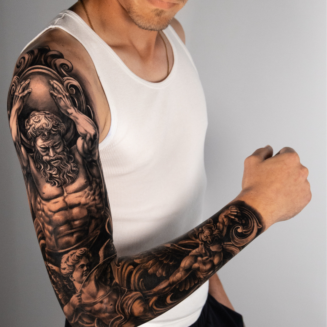Close-up of a man's arm with an intricate black and gray tattoo sleeve featuring mythological figures and swirling patterns.