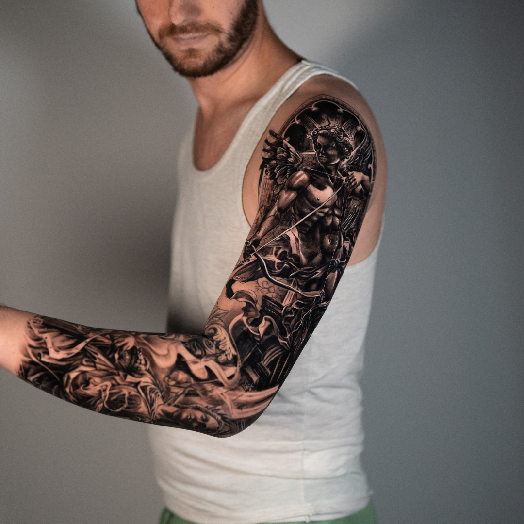 Tattoo of a muscular angel with wings, holding a bow and arrow, on a man's upper arm and shoulder.