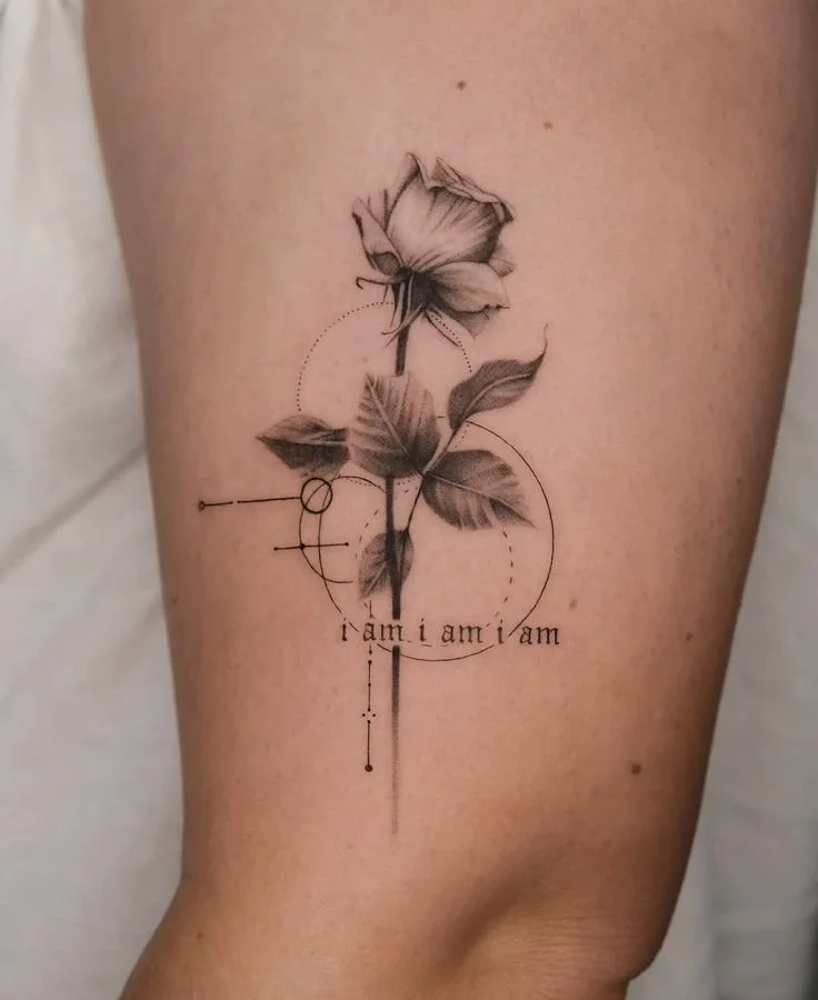 Fine line rose thigh tattoo with geometric accents and “i am” script—minimalist tattoo ideas for women with soft black and grey shading.
