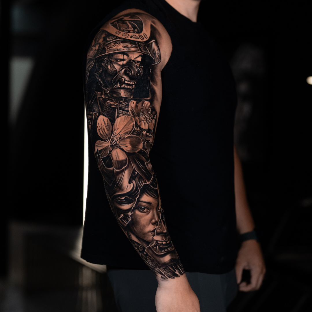Close-up of a person's arm with a detailed black and gray tattoo sleeve featuring a woman's face, flowers, and a soldier wearing a helmet.