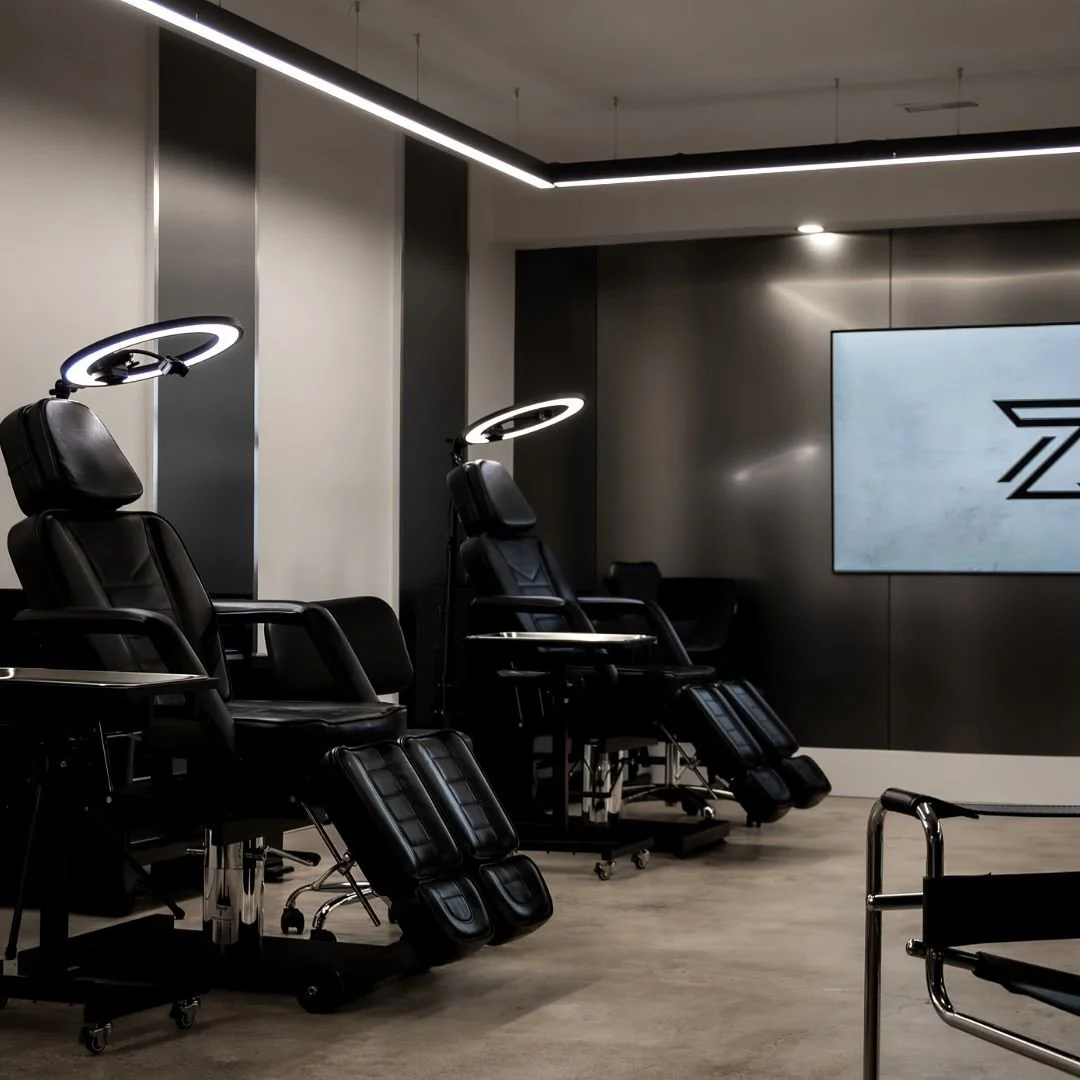 Two black tattoo chairs under ring lights on the artist floor at Zen Tattoo Studio in Oakville, with the Zen Z logo on a wall screen and a chrome framed chair in the foreground.