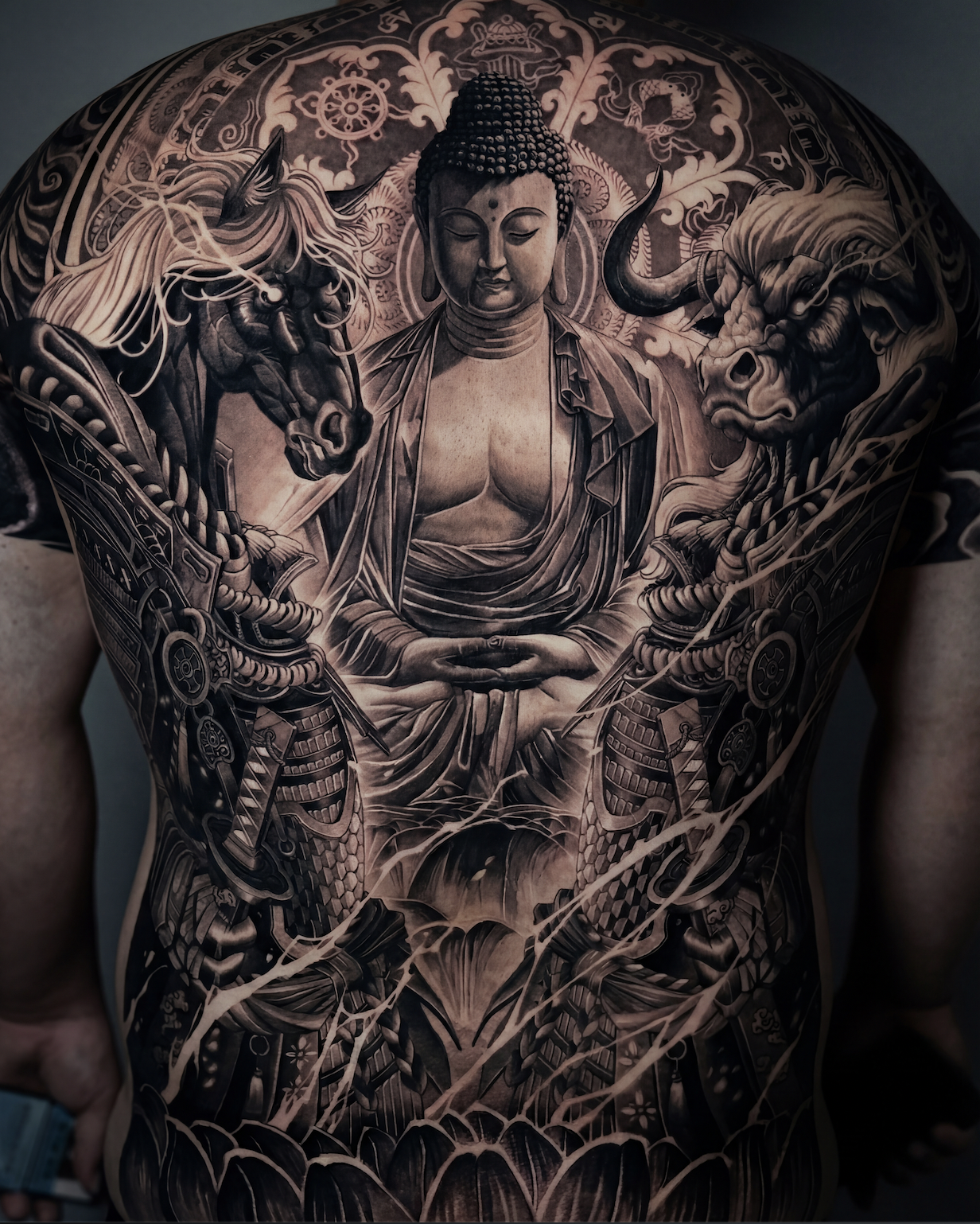Black and grey realism full back tattoo for men with Buddha centerpiece, horse and bull, lotus, and ornamental background