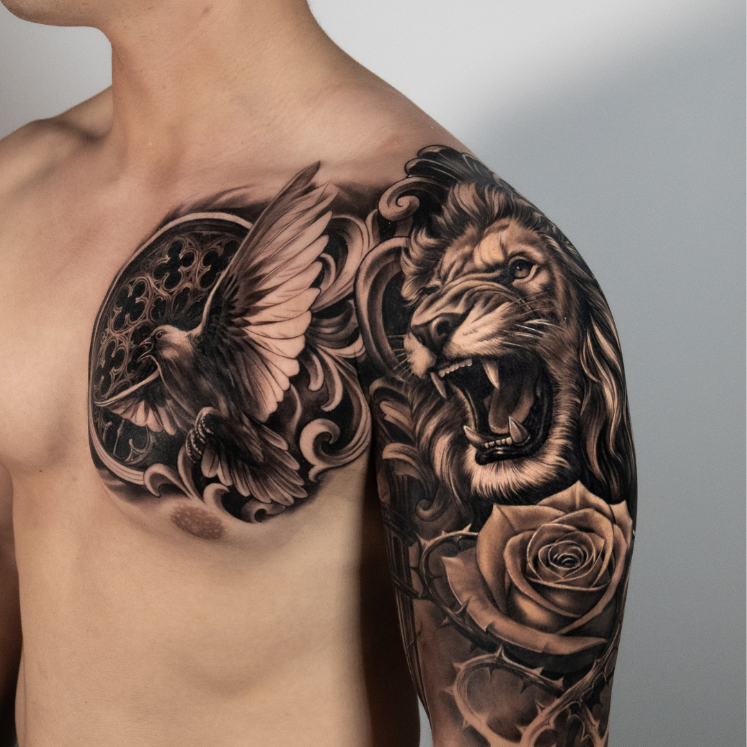 Tattoo of a roaring lion's face alongside a rose bouquet, with a flying bird and decorative elements on a man's shoulder and upper arm.