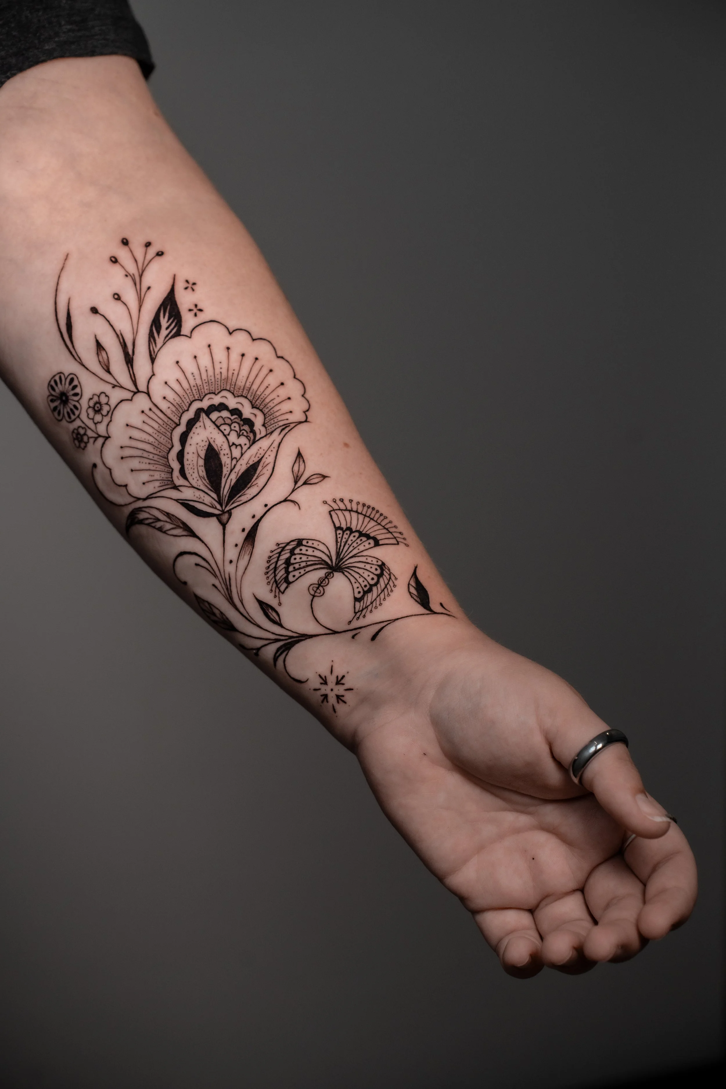 Close-up of a person's arm with a black line art floral and butterfly tattoo, bent at the wrist with hand relaxed, wearing a metallic ring.