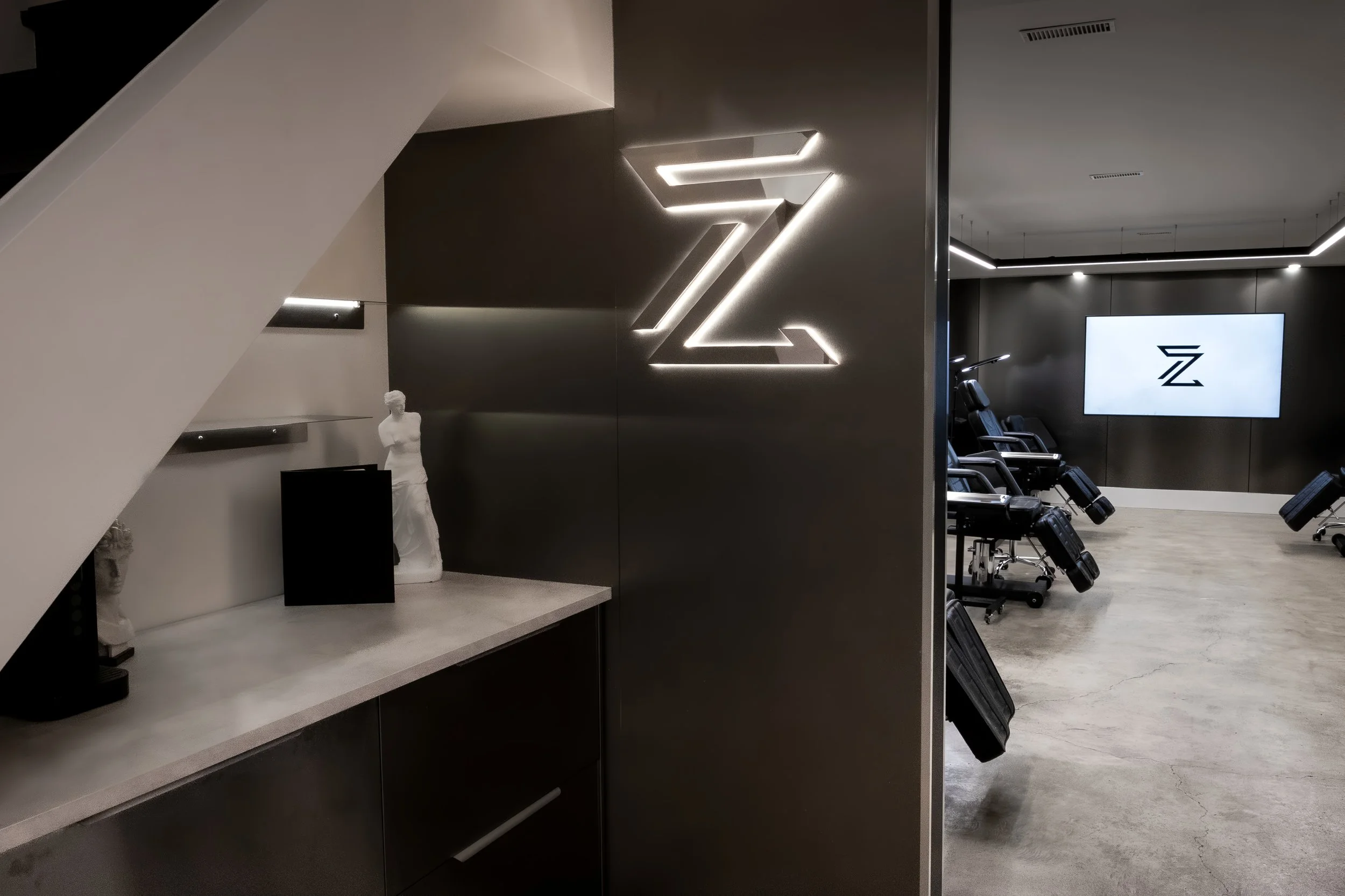 Interior of a modern gym or fitness center with black walls, a large illuminated 'Z' logo on the wall, massage chairs, and a digital screen displaying a 'Z' logo.