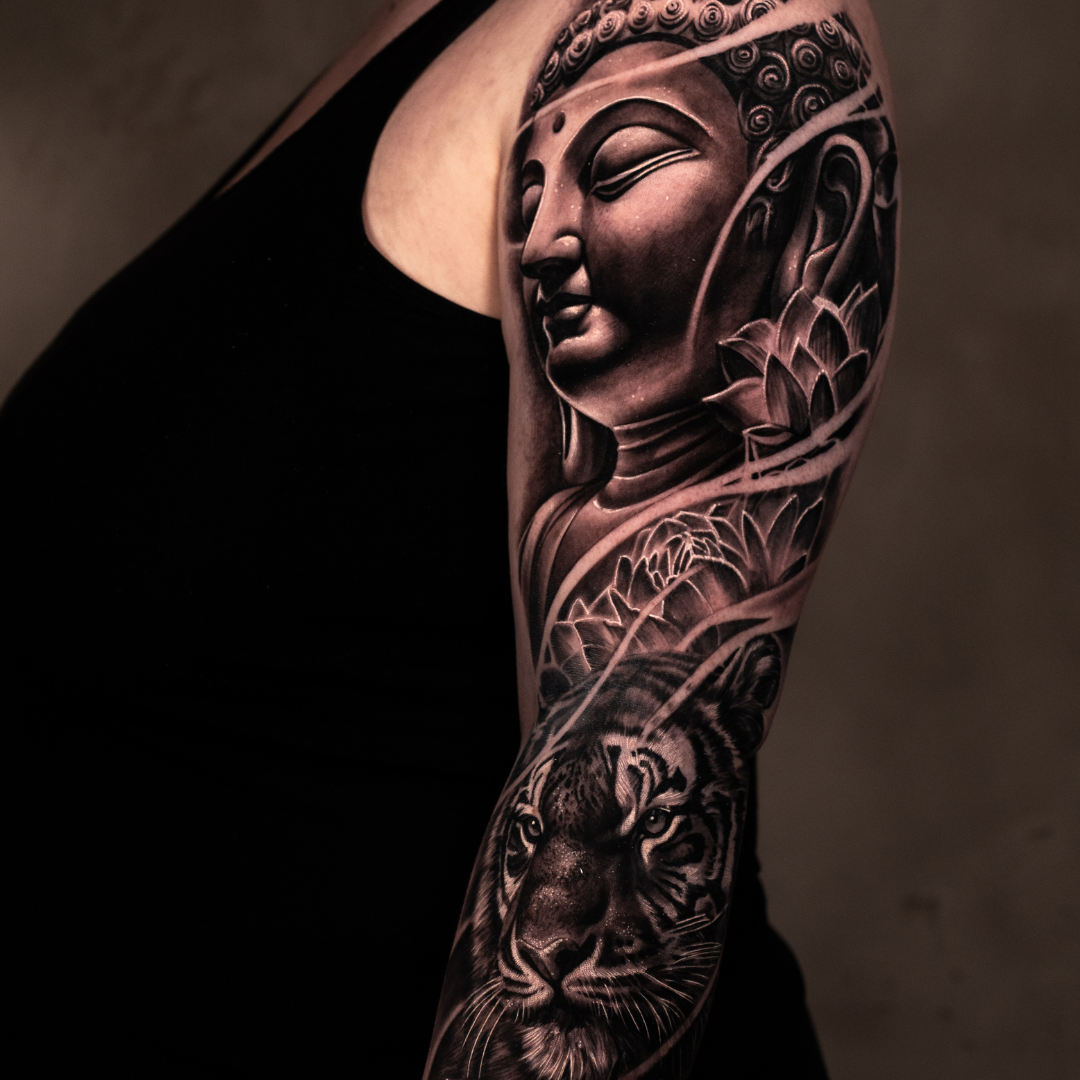 Tattoo of a Buddha's face with closed eyes and serene expression on upper arm, alongside a detailed tiger head tattoo below, with floral and ornamental elements