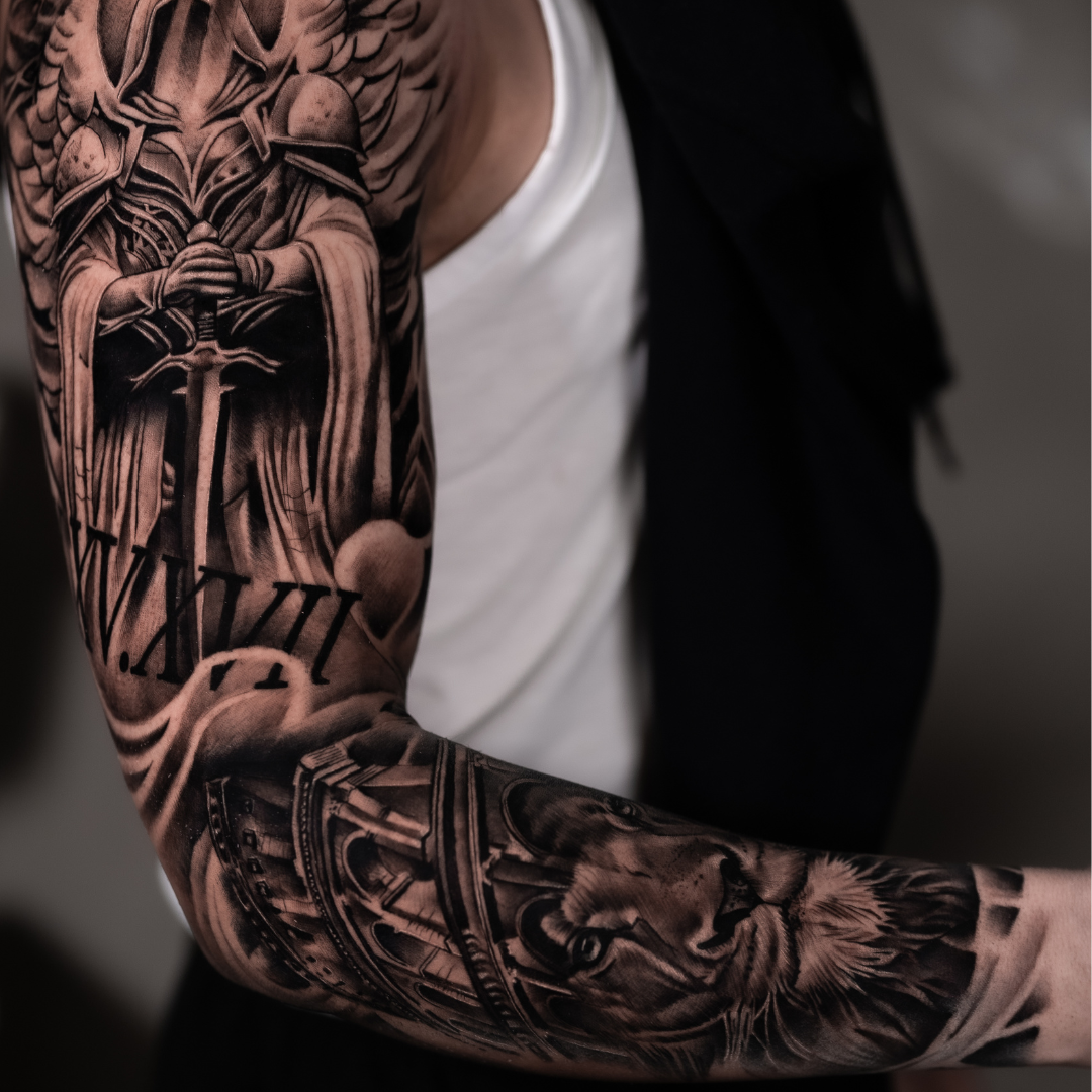 Close-up of a person's arm with detailed black and gray tattoo sleeve, including a sword, Roman numeral XII, a lion, and architectural elements.