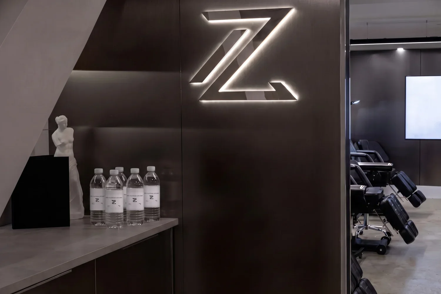 Zen water bottles on the counter by the lit Zen logo wall at Zen Tattoo Studio in Oakville, with the studio's tattoo chairs beyond.