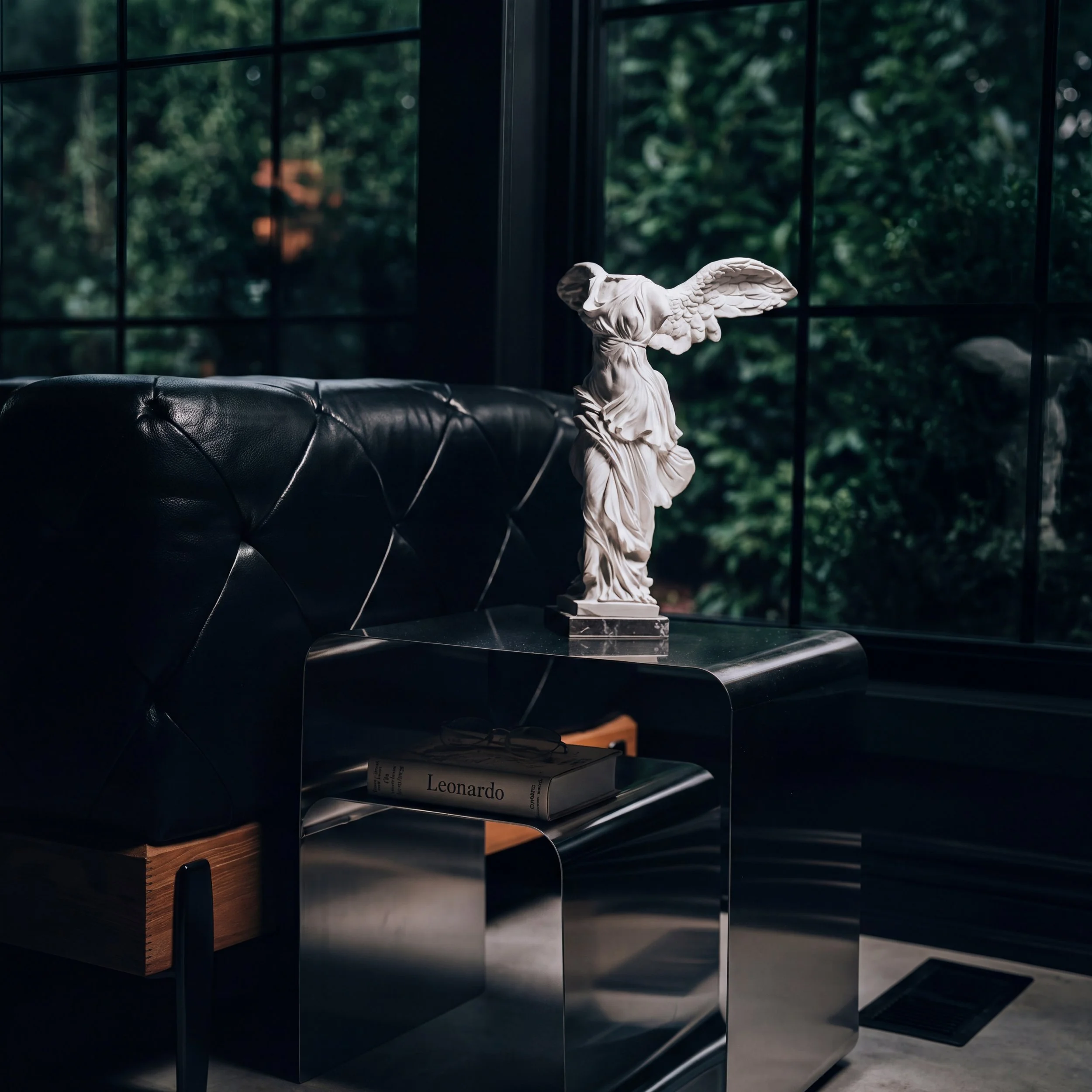 A white Winged Victory statue on a black glass side table beside a tufted black leather sofa, in front of tall dark framed windows at Zen Tattoo Studio in Oakville.