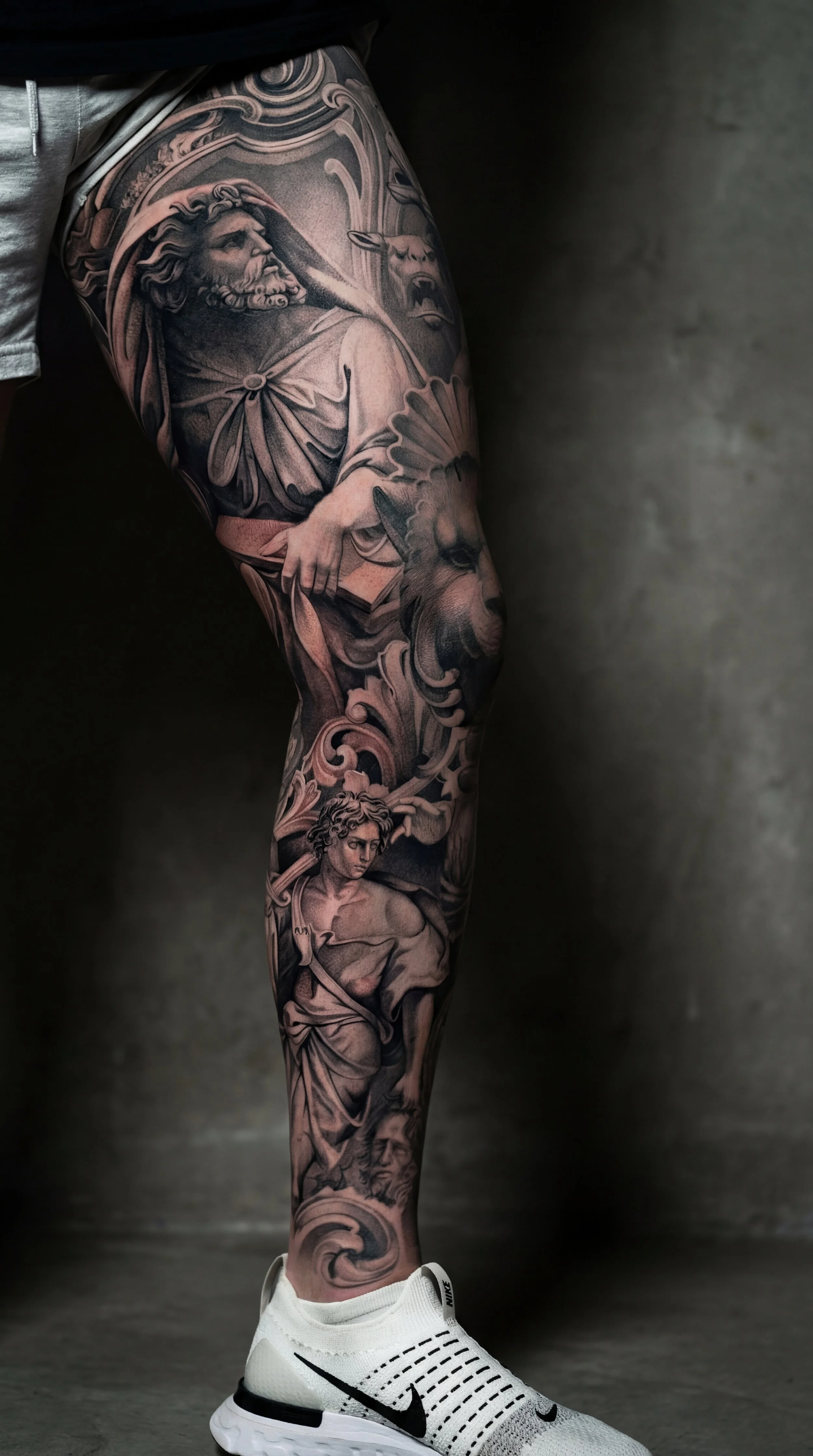 Leg tattoo ideas for guys header image featuring a black and grey realism leg sleeve with Greek statue style figures and detailed shading across the thigh calf and shin