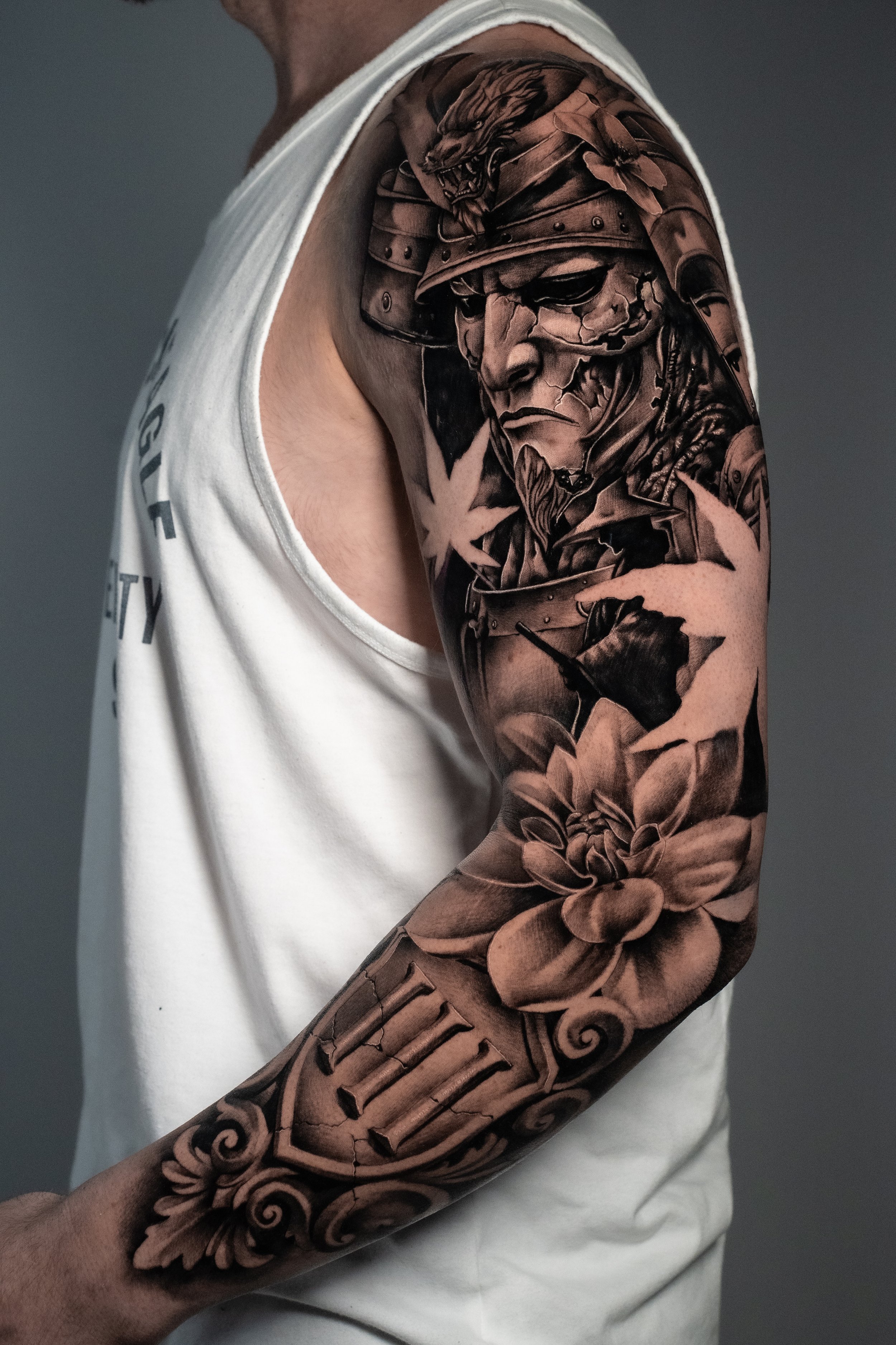 A detailed black and gray tattoo sleeve featuring a samurai warrior with a fierce expression, wearing a helmet and armor, with floral and ornamental designs surrounding the figure on the person's arm.