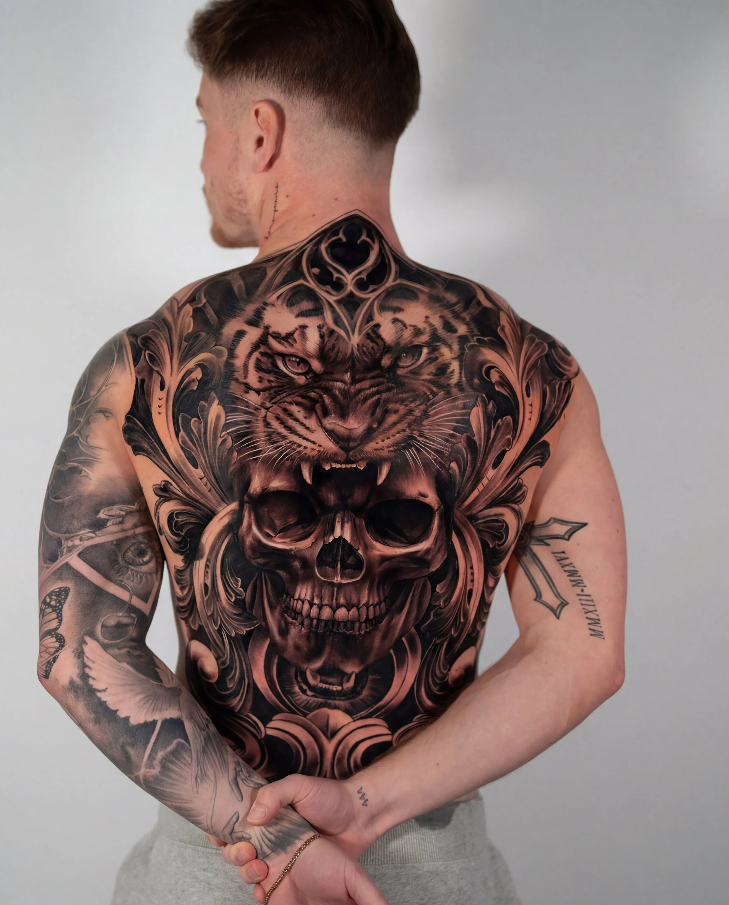 Alt text: Back tattoo for men with tiger head above a skull and ornamental framing in bold black and grey realism