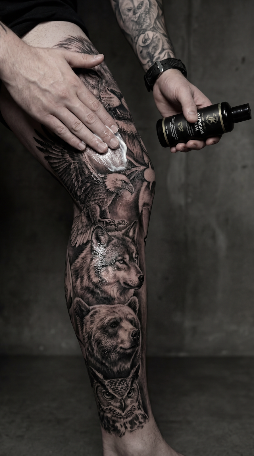Leg tattoo ideas for guys showing a black and grey realism leg sleeve with wolf and bear portraits eagle and owl details while applying tattoo aftercare cream during healing