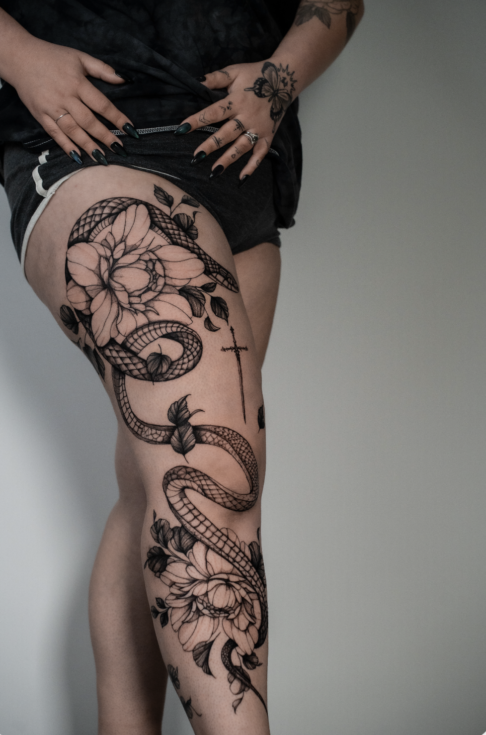 A person with black tattoos, including a snake, flowers, a cross, and a butterfly, on their thigh. They are lifting their shirt slightly and have dark painted nails.