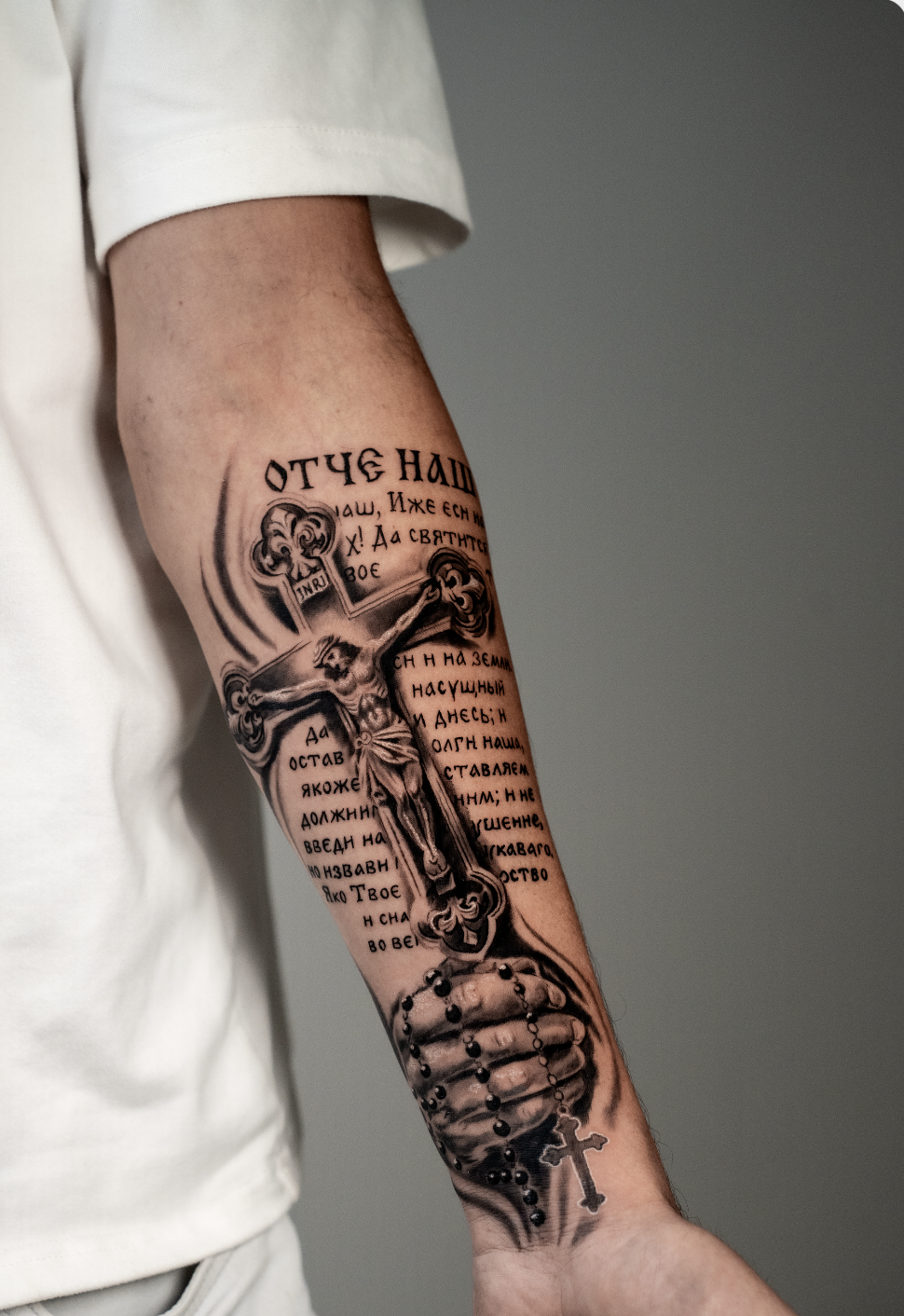 Tattoo of Jesus on the cross with Russian text and a rosary on a person's forearm.