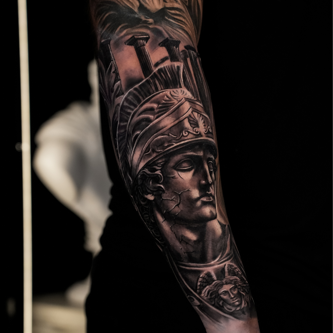 A close-up of a tattoo sleeve on a person's arm, featuring a detailed grayscale portrait of a classical warrior wearing a helmet, with other intricate designs.