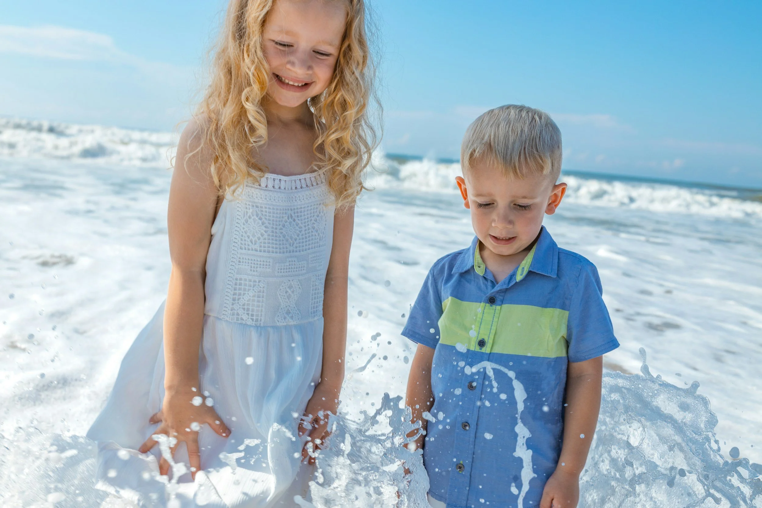 A young girl with curly blonde hair wearing a white dress and a young boy with short blonde hair wearing a blue and green shirt standing in the ocean with waves and blue sky behind them.