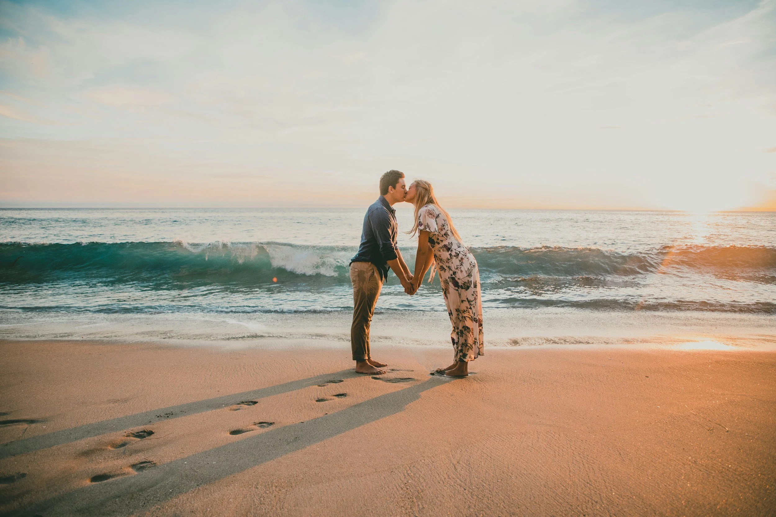 A couple holding hands and kissing on the beach at sunset, with footprints in the sand and the ocean waves in the background.