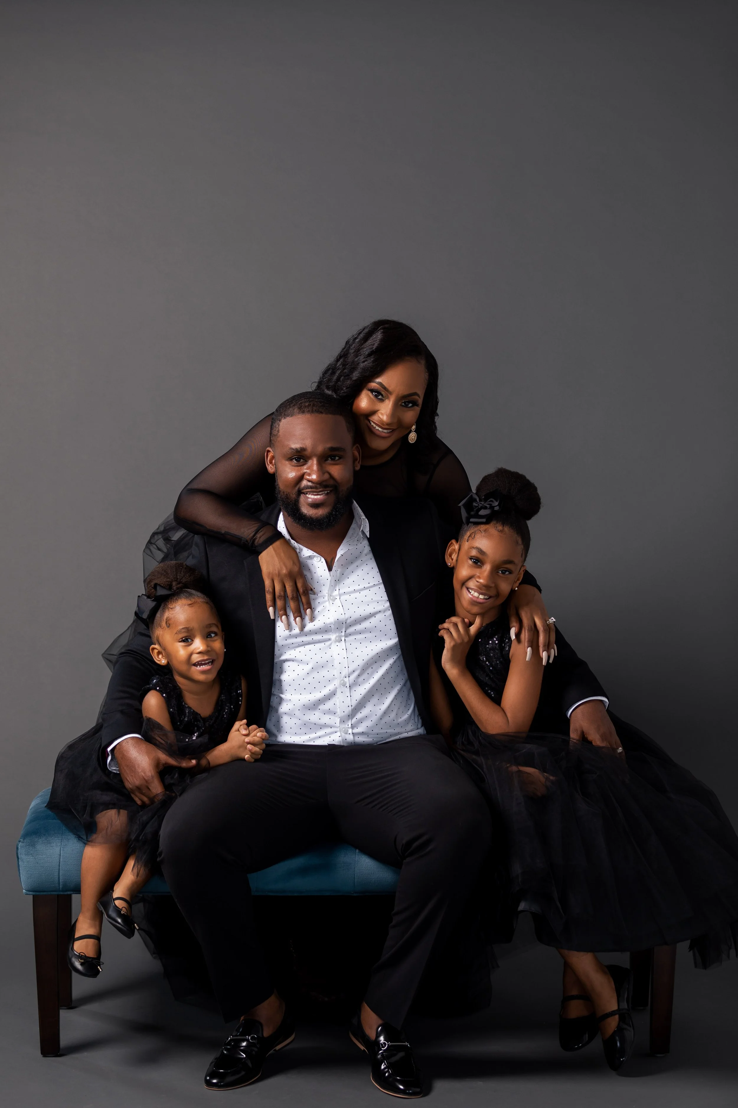 A smiling family of five posing together against a gray background, with the parents seated on a bench and the children and mother standing around them. The family appears to be dressed in formal black attire.