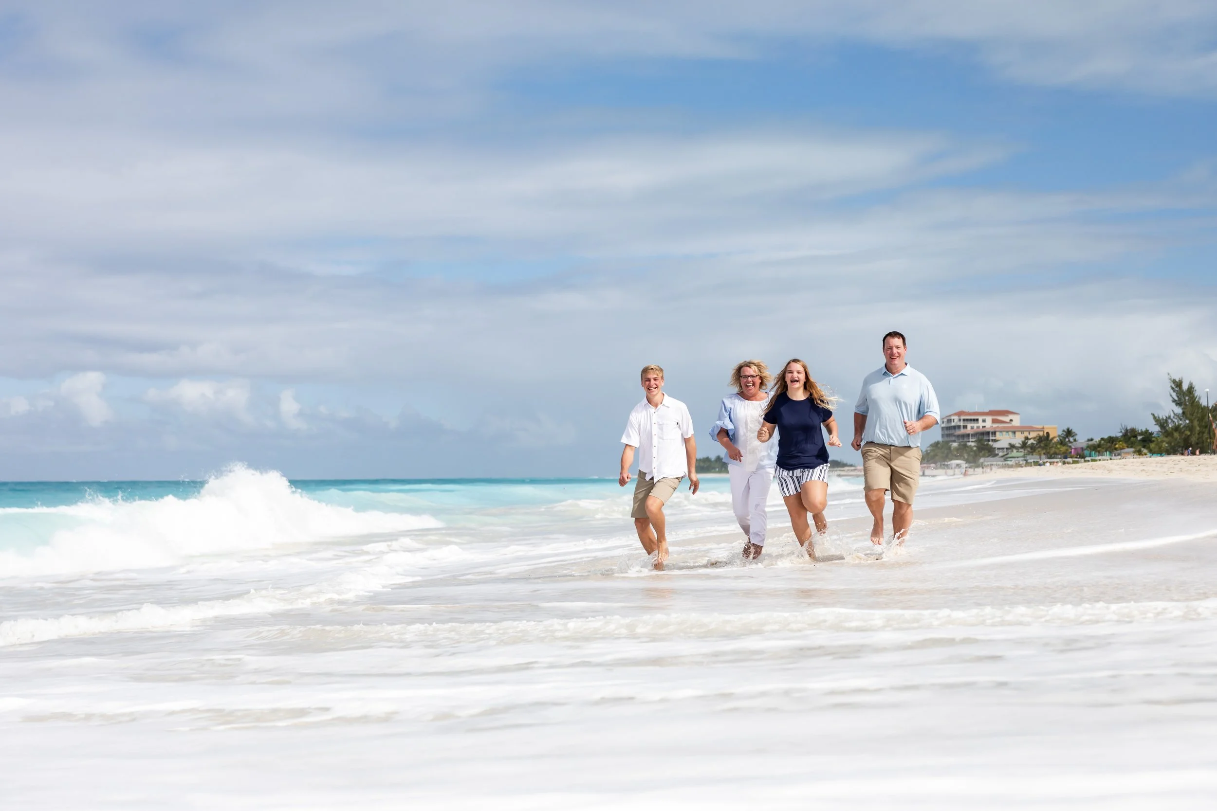 Four smiling people walking and running along the shoreline at the beach with waves, sandy beach, and buildings in the background under a partly cloudy sky.