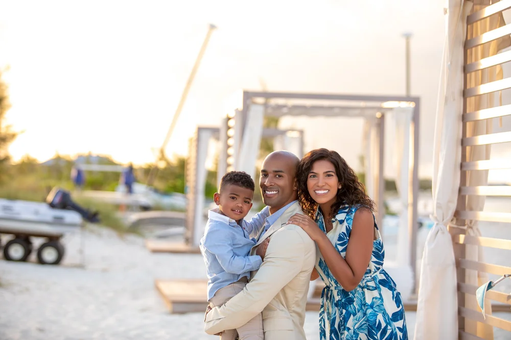 A smiling family of three, including a man, woman, and young boy, standing on a beach near a wooden cabana during sunset.