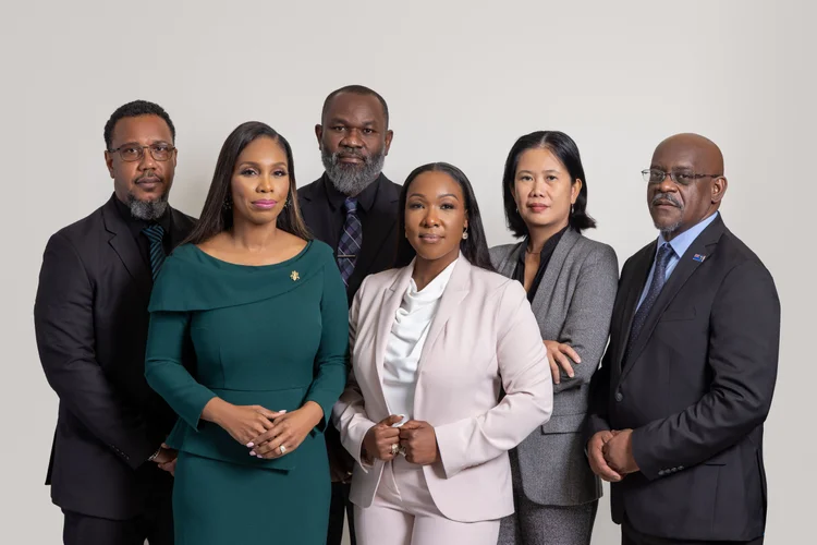 Six diverse professionals dressed in business attire posing for a group photo against a plain light gray background.