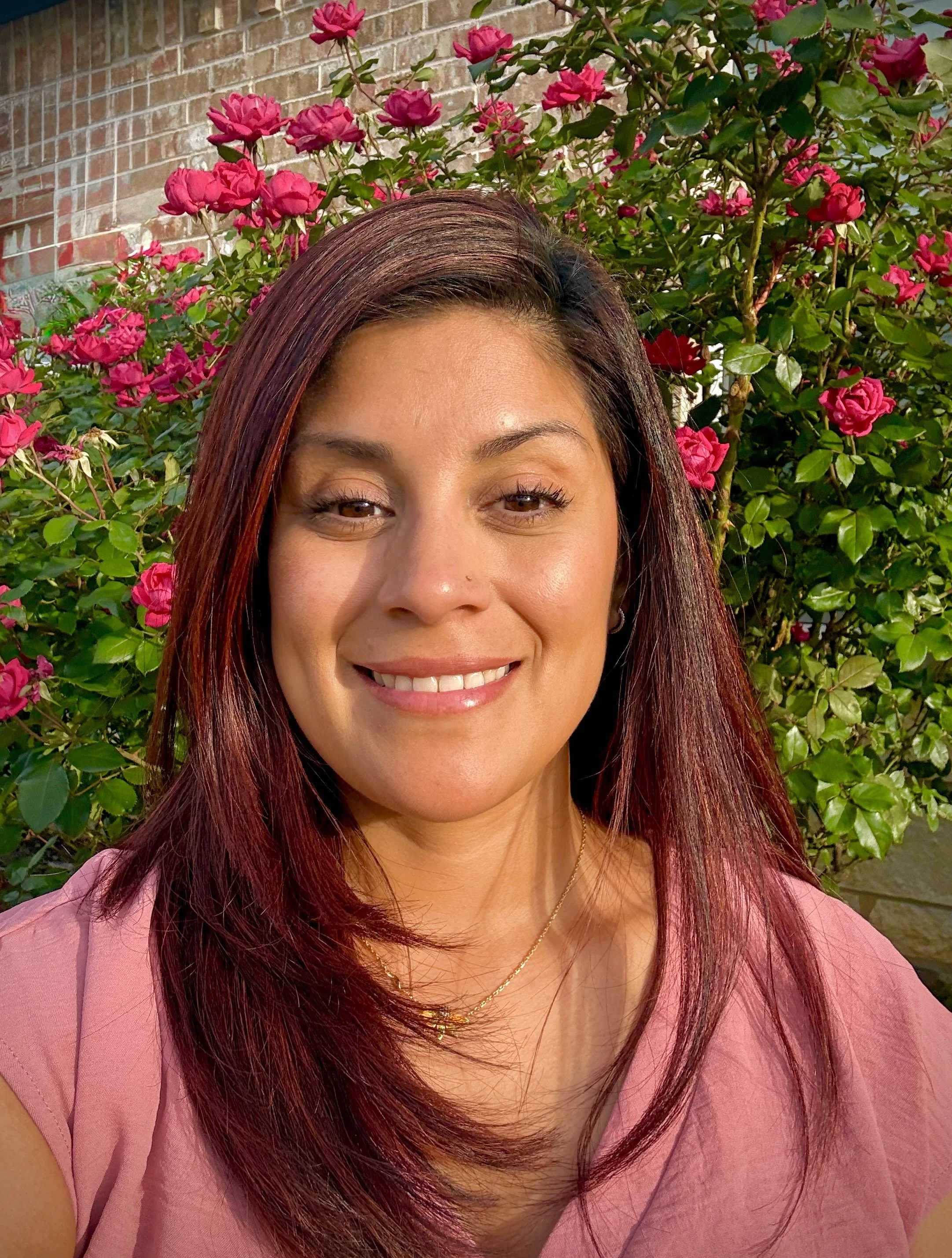 Close-up of a young woman with long, reddish hair, brown eyes, and tan skin, smiling outdoors with a rosebush in the background.
