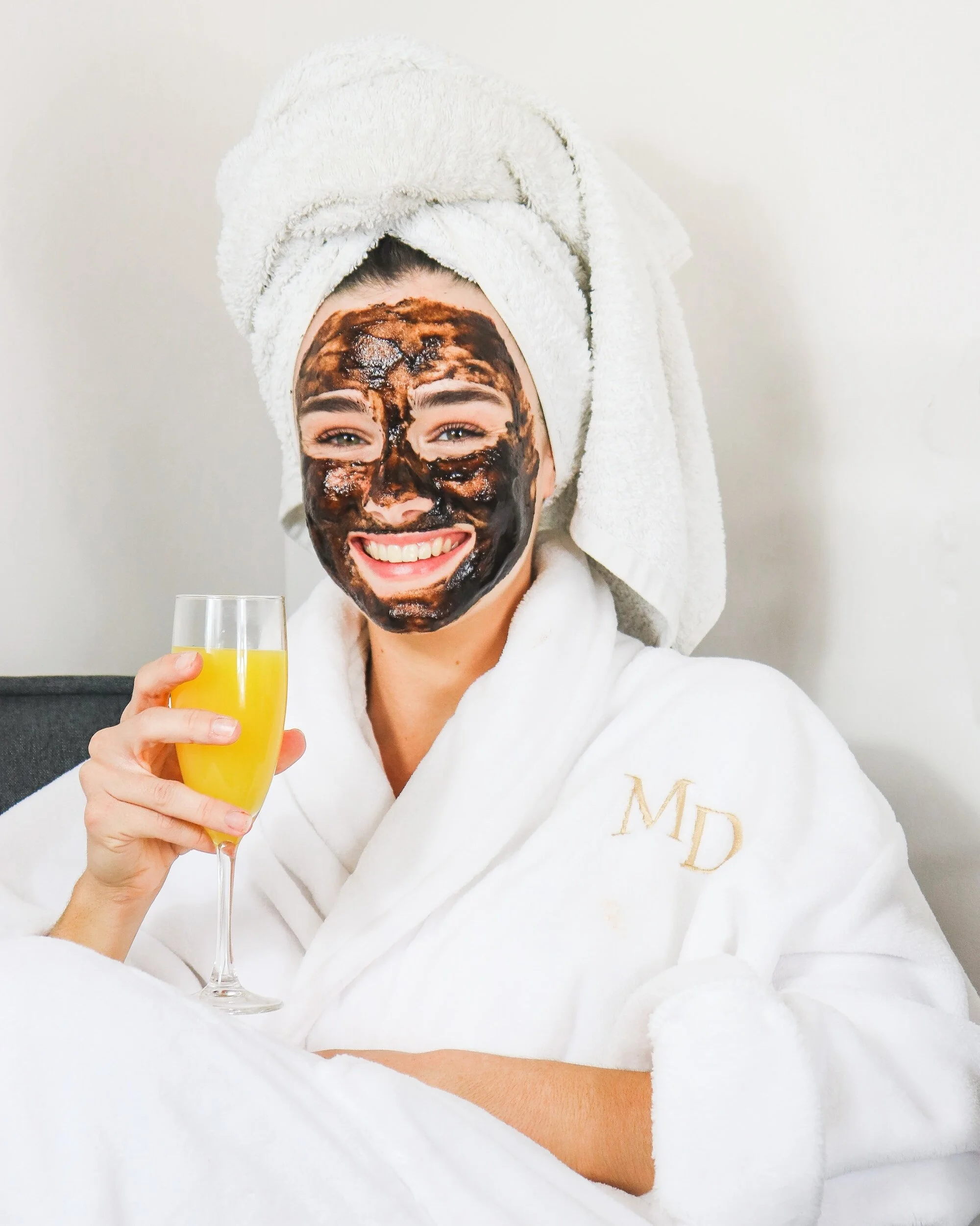 A woman wrapped in a cozy robe, with a mud mask on her face, and mimosa in hand