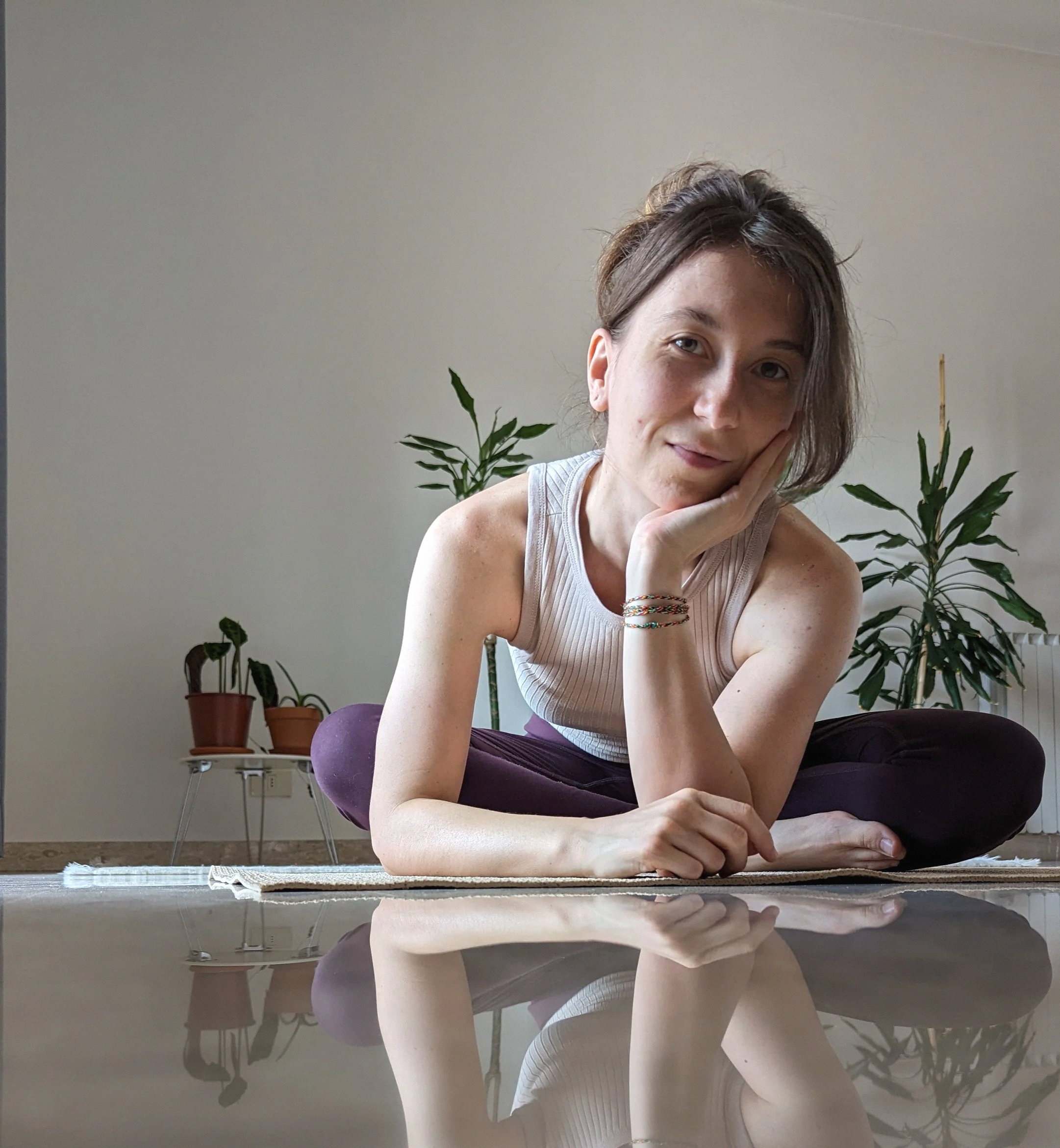 Una donna in una posa rilassata, seduta a terra con una gamba piegata, guarda verso la fotocamera, in un ambiente domestico con piante verdi sullo sfondo.