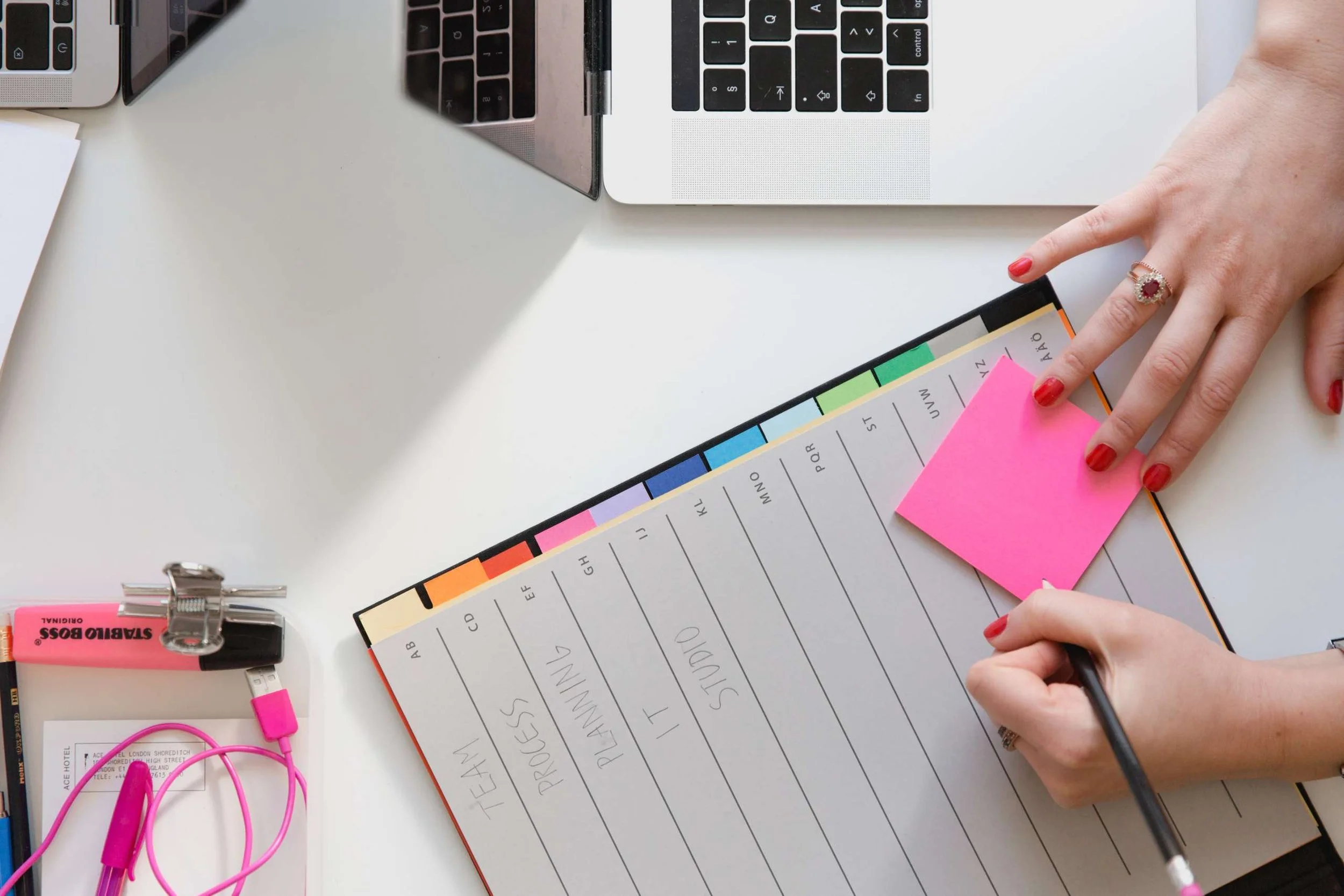 Overhead view of a white desk with a person's hand writing on a planner or notebook, colored sticky tabs on the pages, a laptop, pink earbuds, and a clipboard with a pink highlighter.