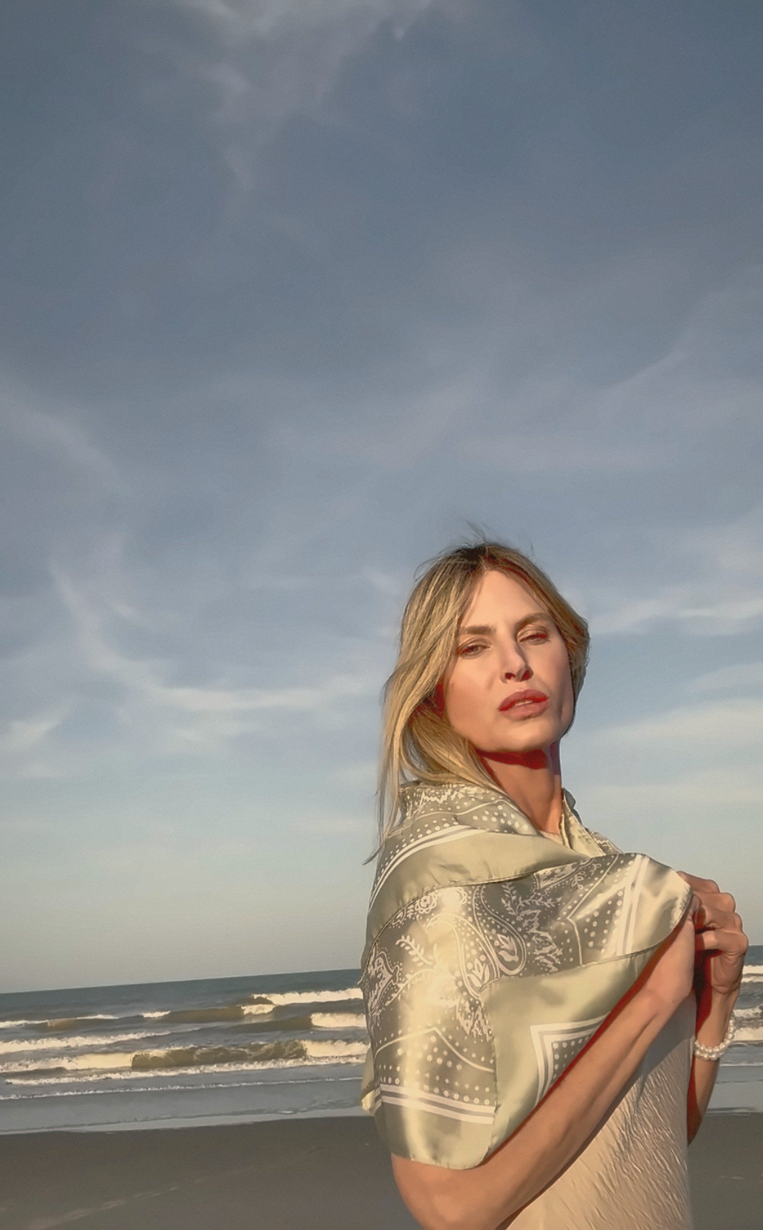 Paige on the beach in a cream dress, a sage silk scarf wrapped over her shoulders, sea and open sky behind her