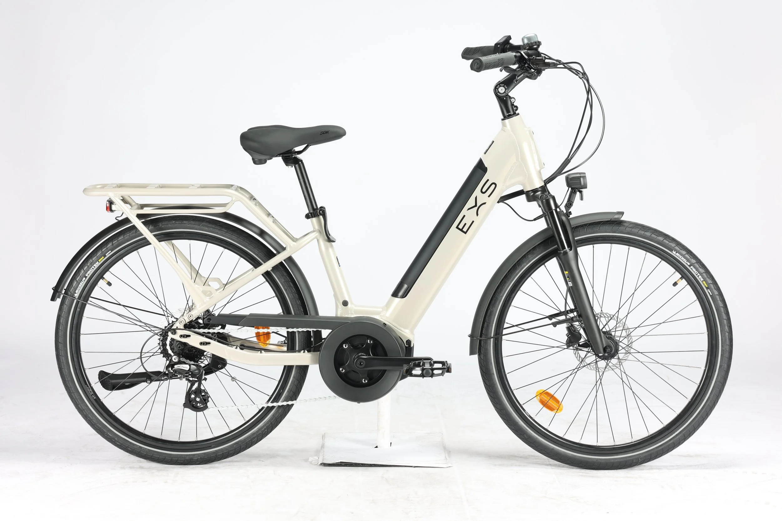 Un vélo électrique blanc avec un cadre moderne, un moteur central, des roues de 26 pouces, des freins à disque, et un porte-bagages arrière. Le vélo est fixé à un support d'exposition contre un fond blanc.