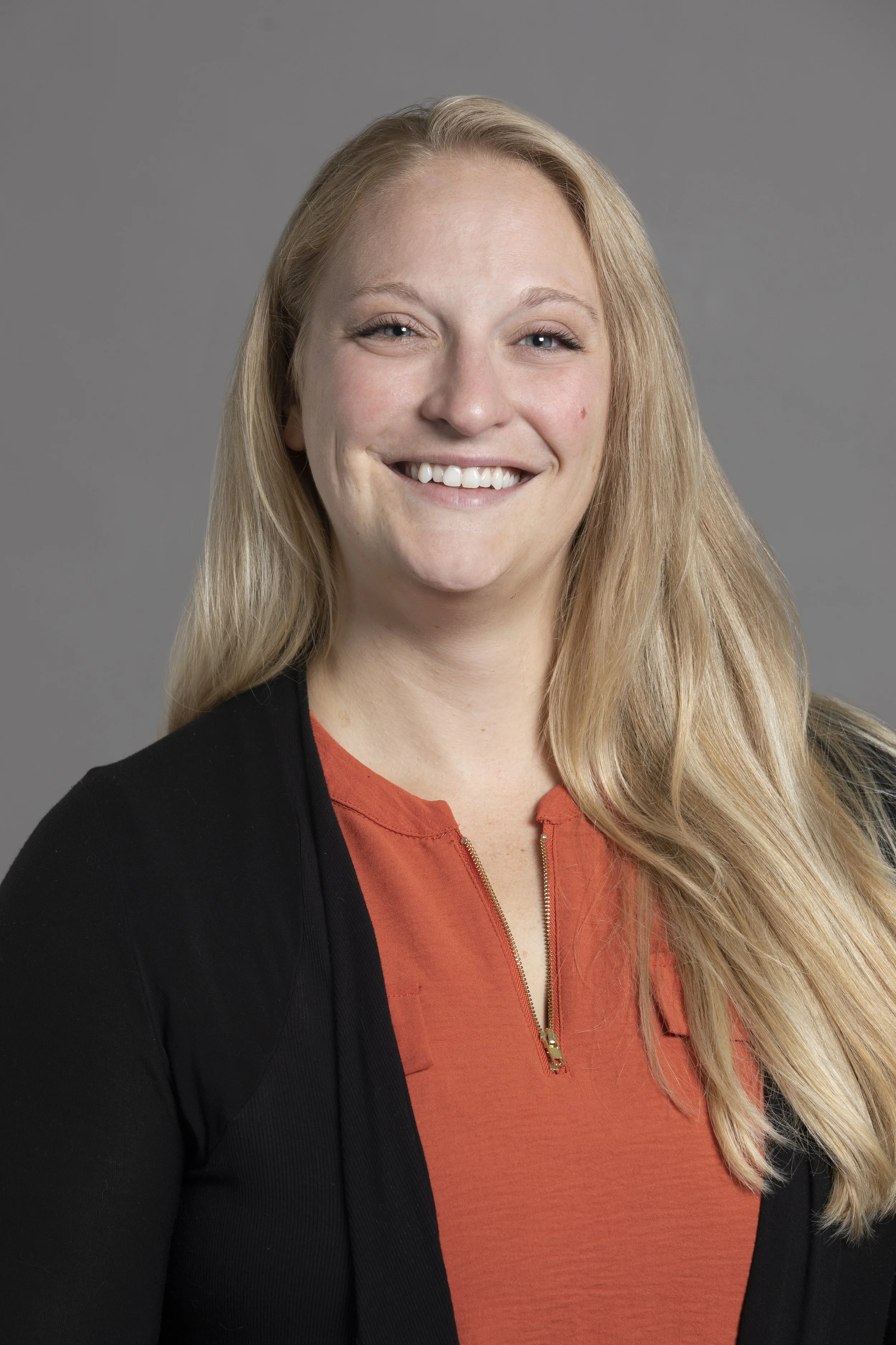 A smiling woman with long blonde hair wearing a black blazer over an orange top with a gold zipper.