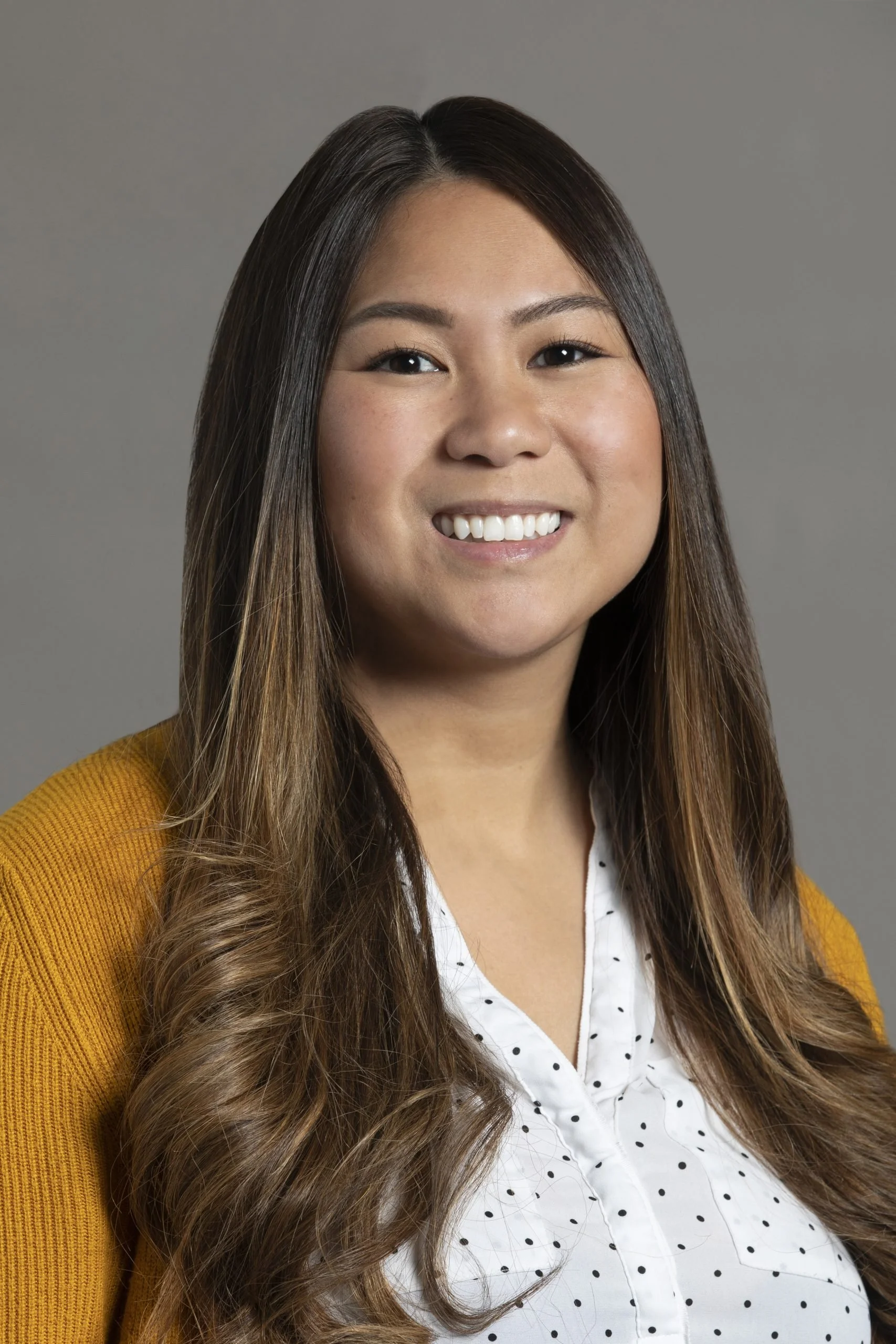 A portrait of a smiling woman with long brown hair, wearing a yellow cardigan over a white shirt with black polka dots, against a plain gray background.