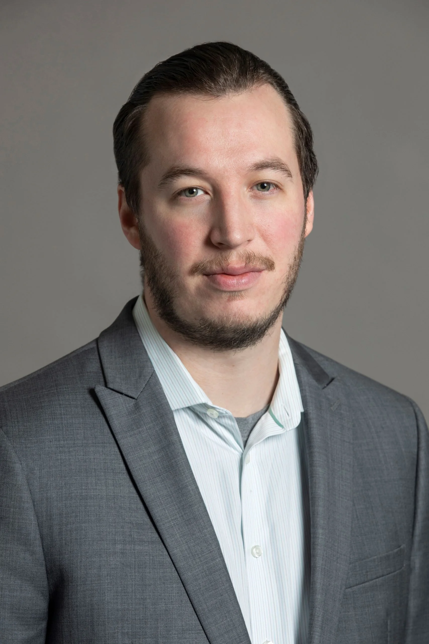 Portrait of a young man with medium-length dark hair, a light beard, wearing a gray suit jacket and a white collared shirt, against a plain gray background.