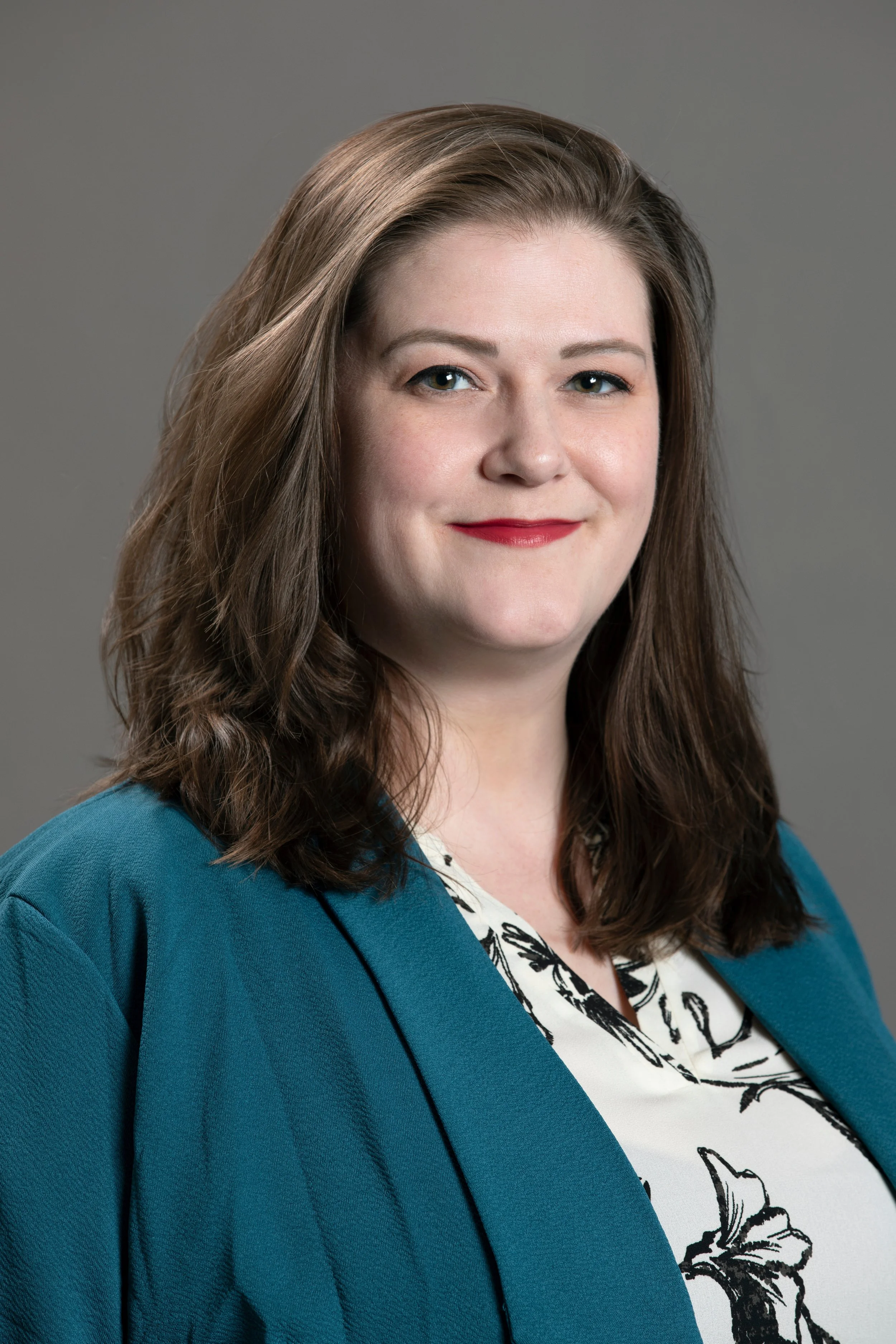 A woman with shoulder-length brown hair, wearing a teal blazer over a white blouse with black floral print, smiling while facing the camera against a plain gray background.