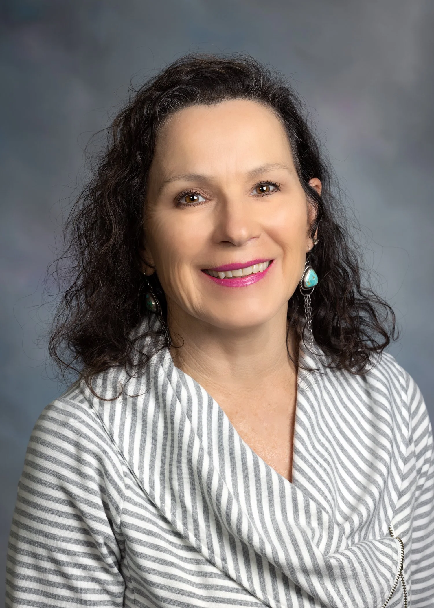 A woman with dark curly hair, wearing a white and gray striped top, smiling, with turquoise earrings and pink lipstick, posed against a gray background.