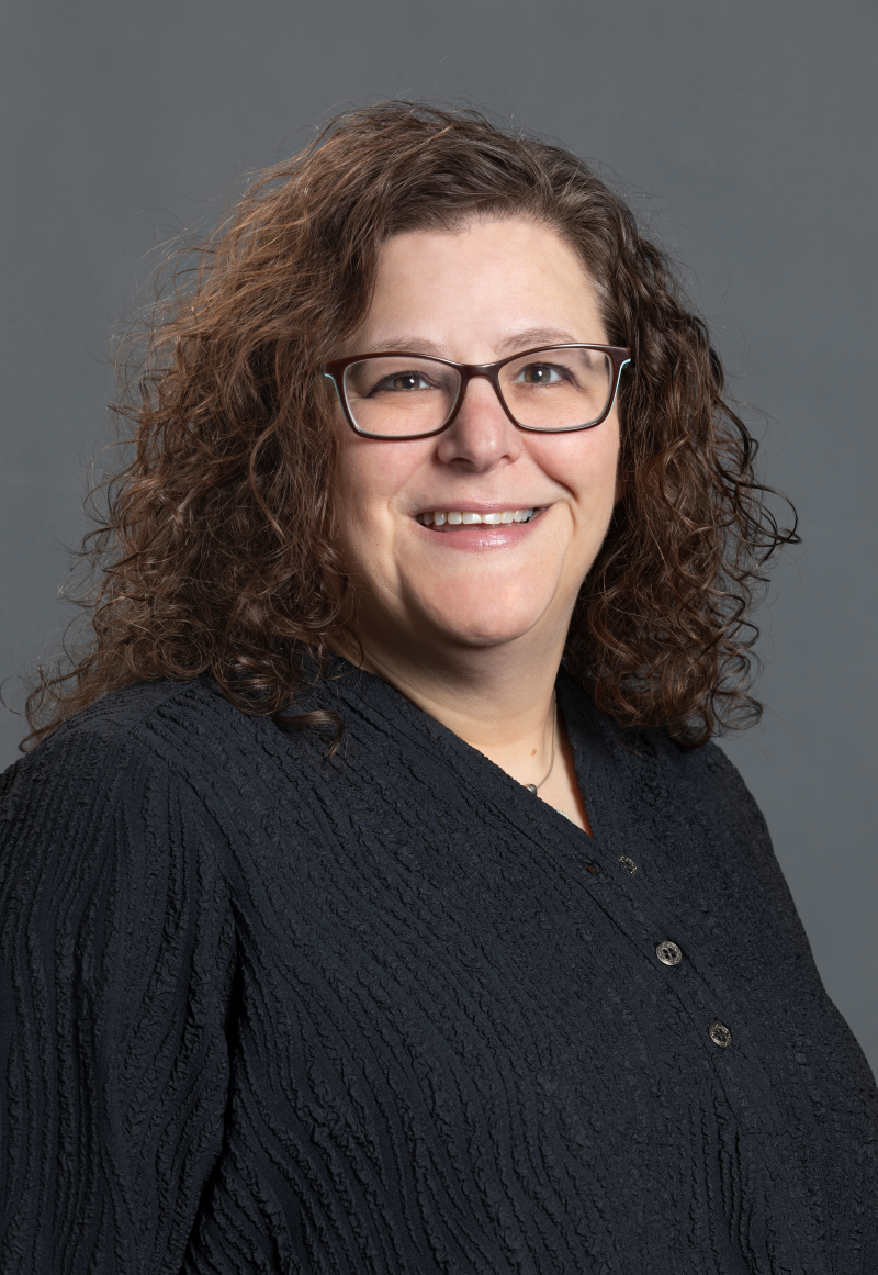 Close-up portrait of a woman with curly brown hair, wearing glasses and a black textured top, smiling against a gray background.