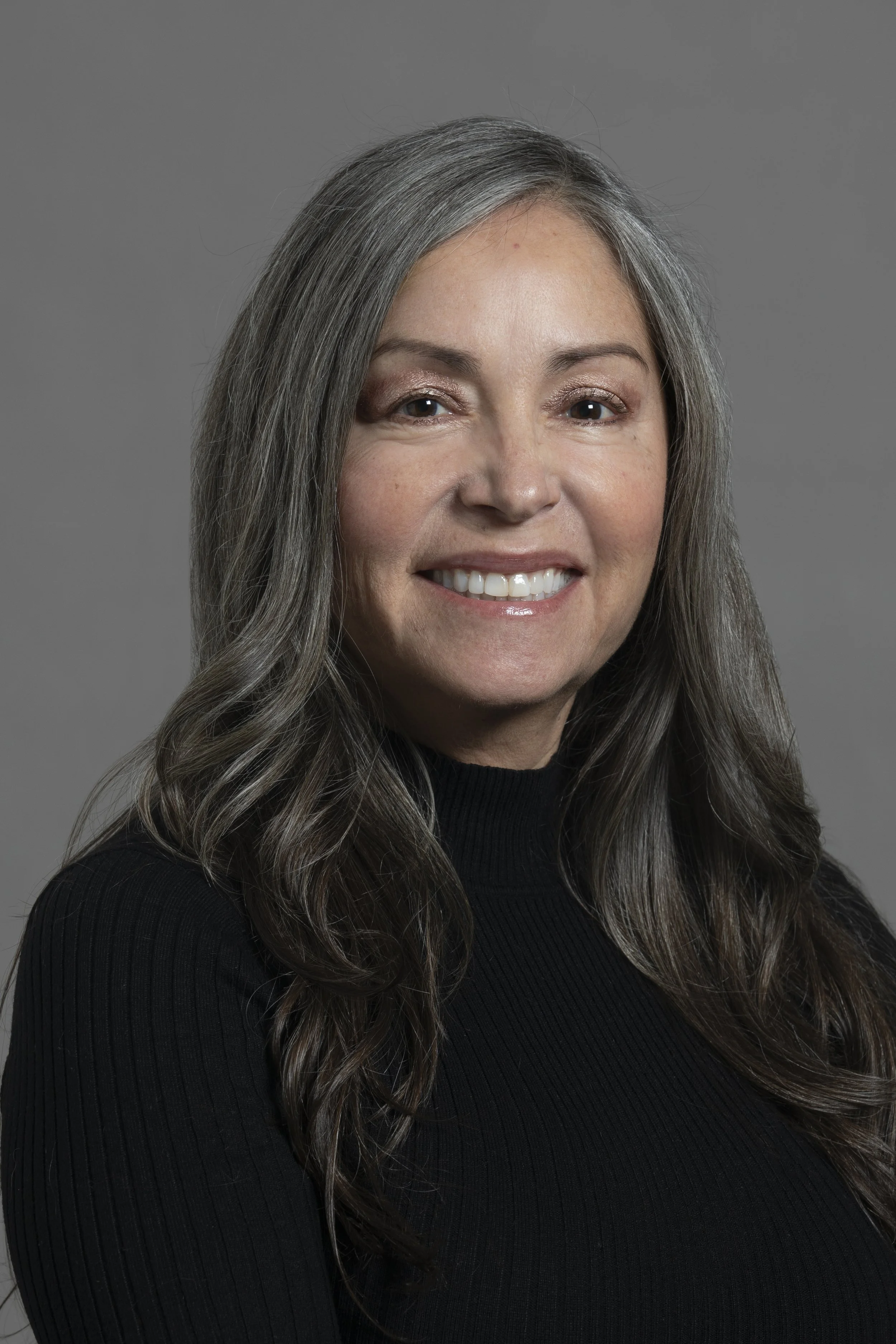 Portrait of a middle-aged woman with long gray hair, smiling, wearing a black top against a gray background.