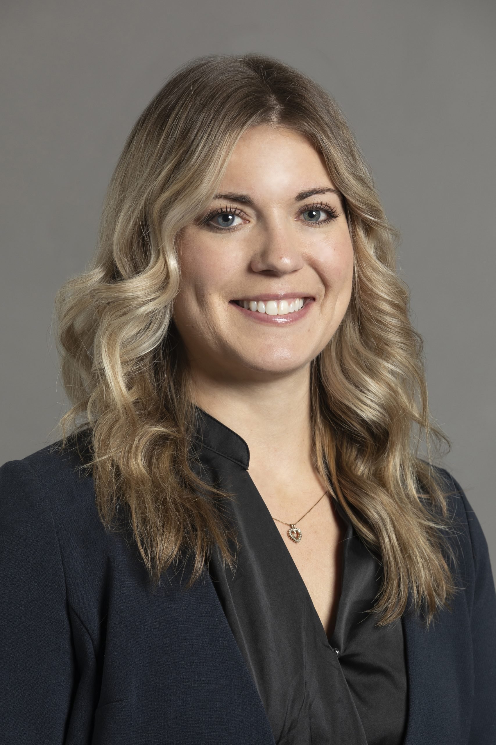 A young woman with wavy blonde hair and blue eyes, wearing a black blazer and a pendant necklace, smiling at the camera against a gray background.