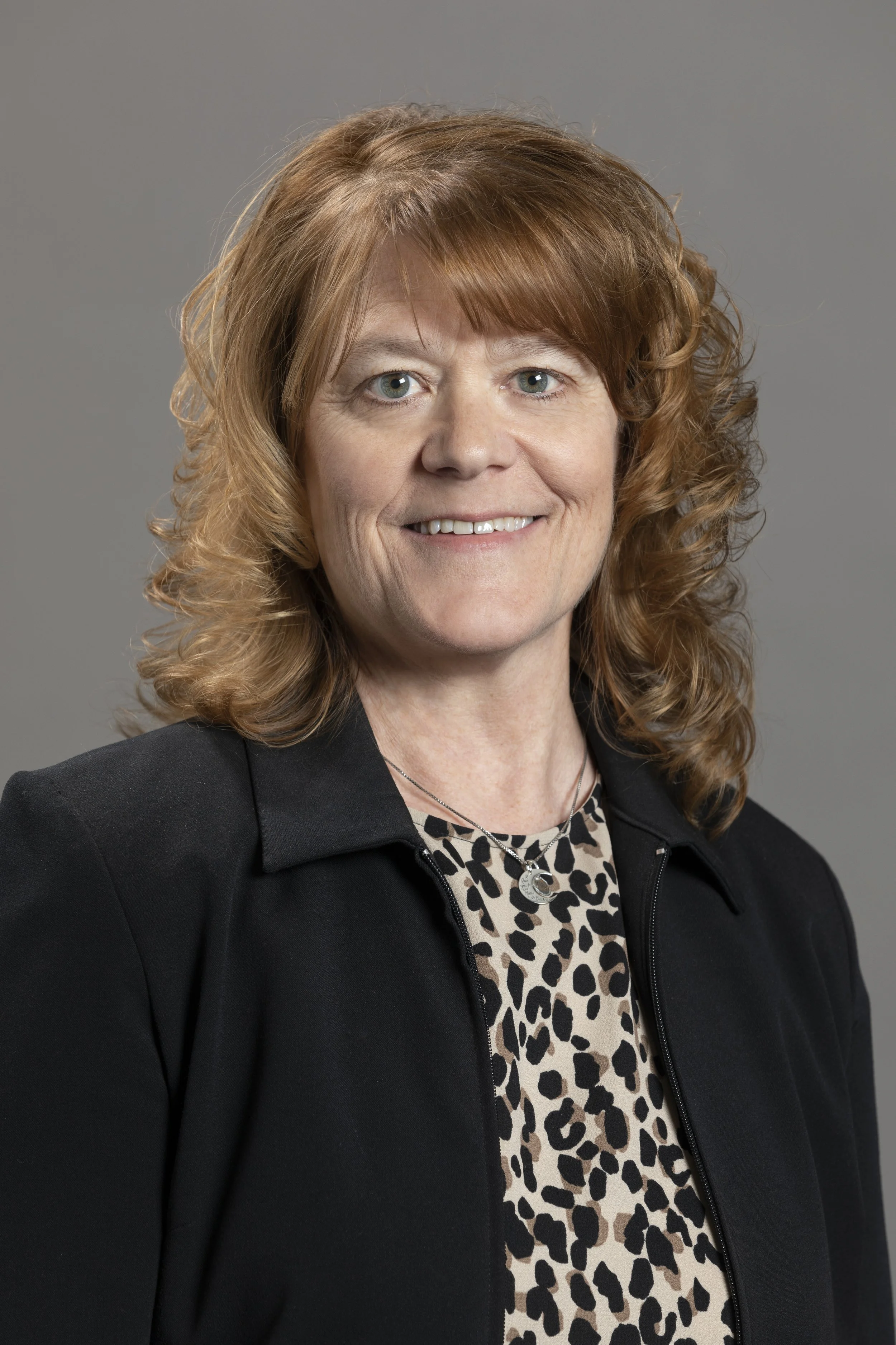 A professional woman with red hair, wearing a black blazer and a leopard print top, smiling against a gray background.