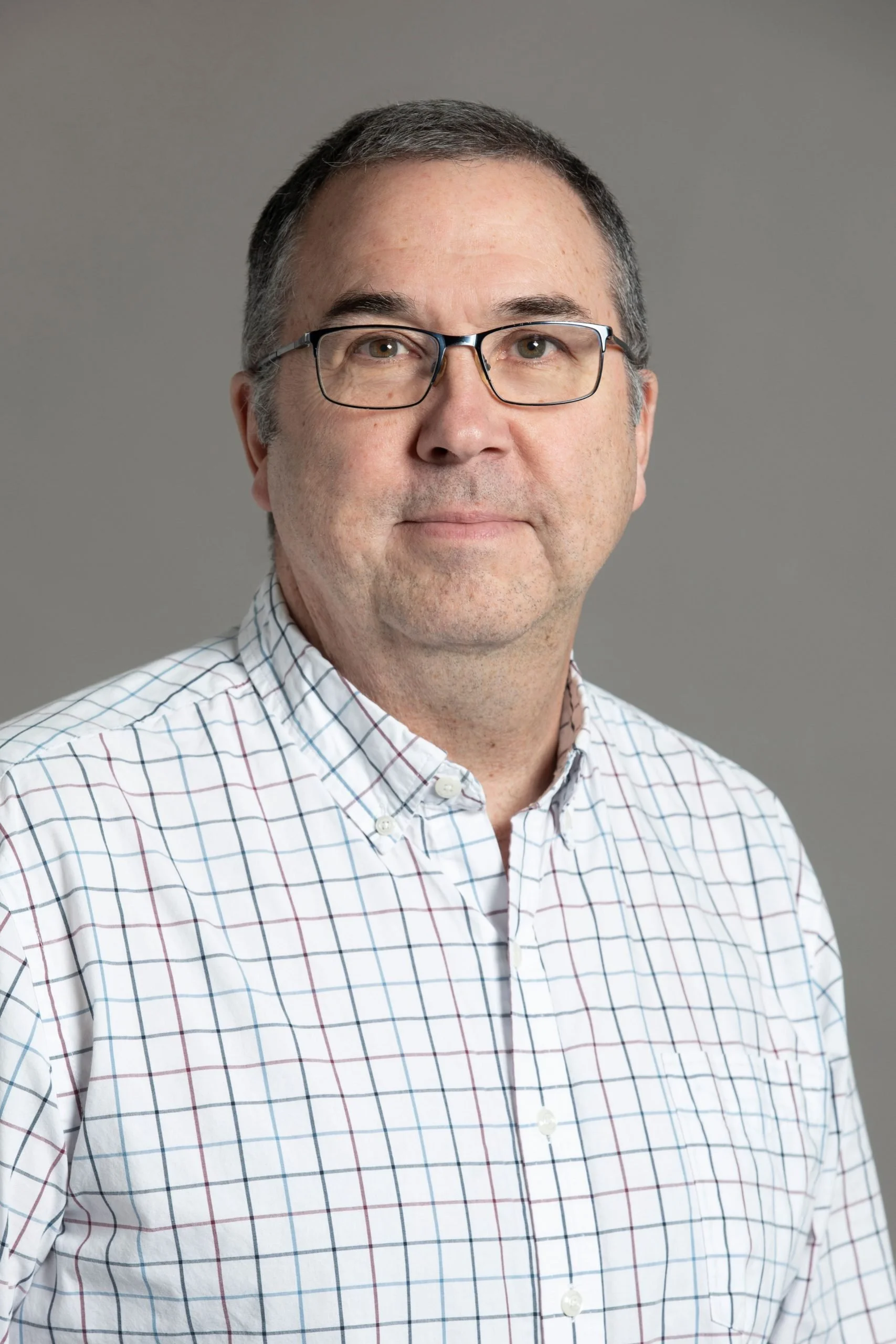 Portrait of a middle-aged man with short dark hair, glasses, wearing a white button-up shirt with a red and blue checkered pattern, standing against a plain gray background.