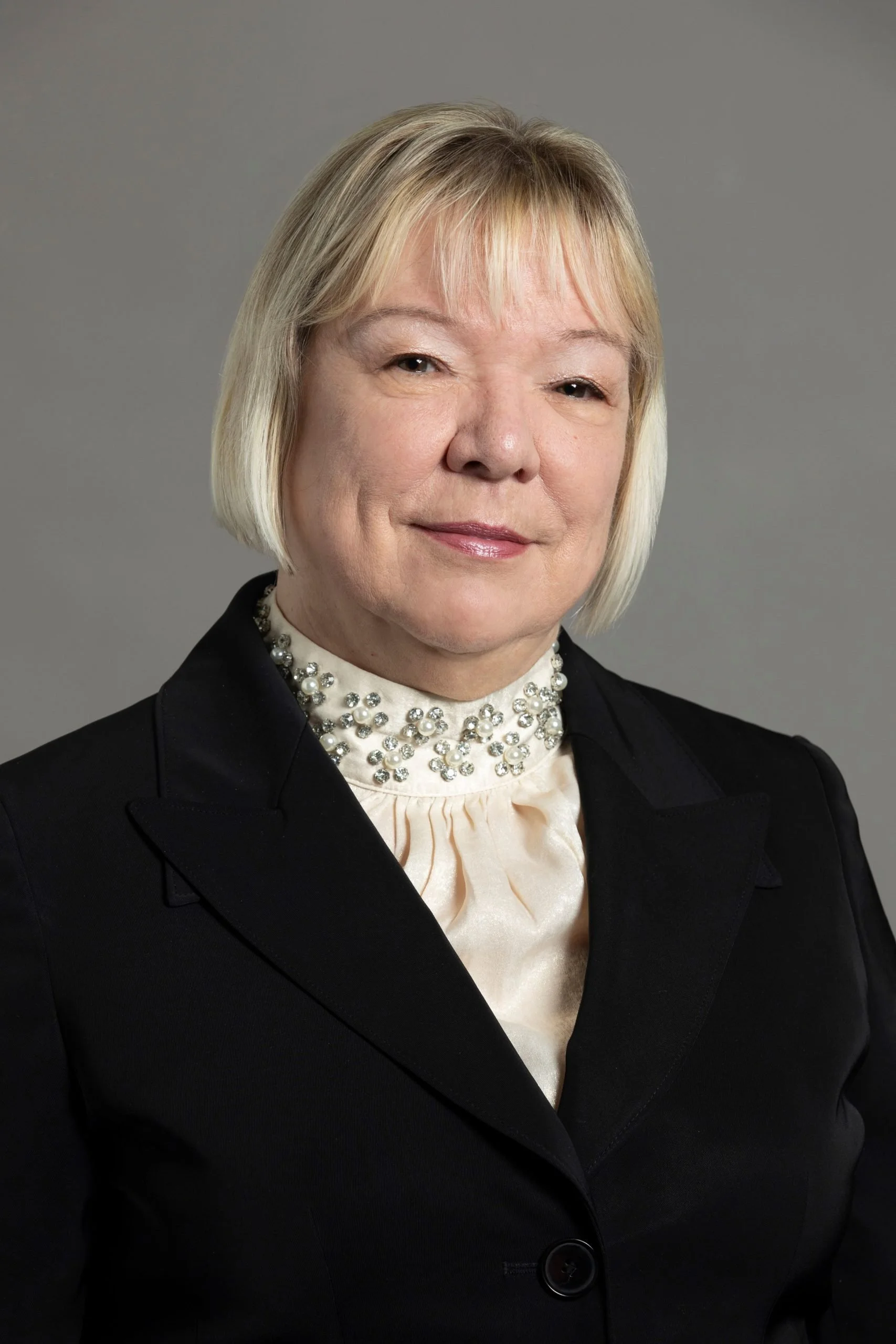 A middle-aged woman with short blonde hair, wearing a black blazer and a white blouse with pearl embellishments, posing against a neutral gray background.