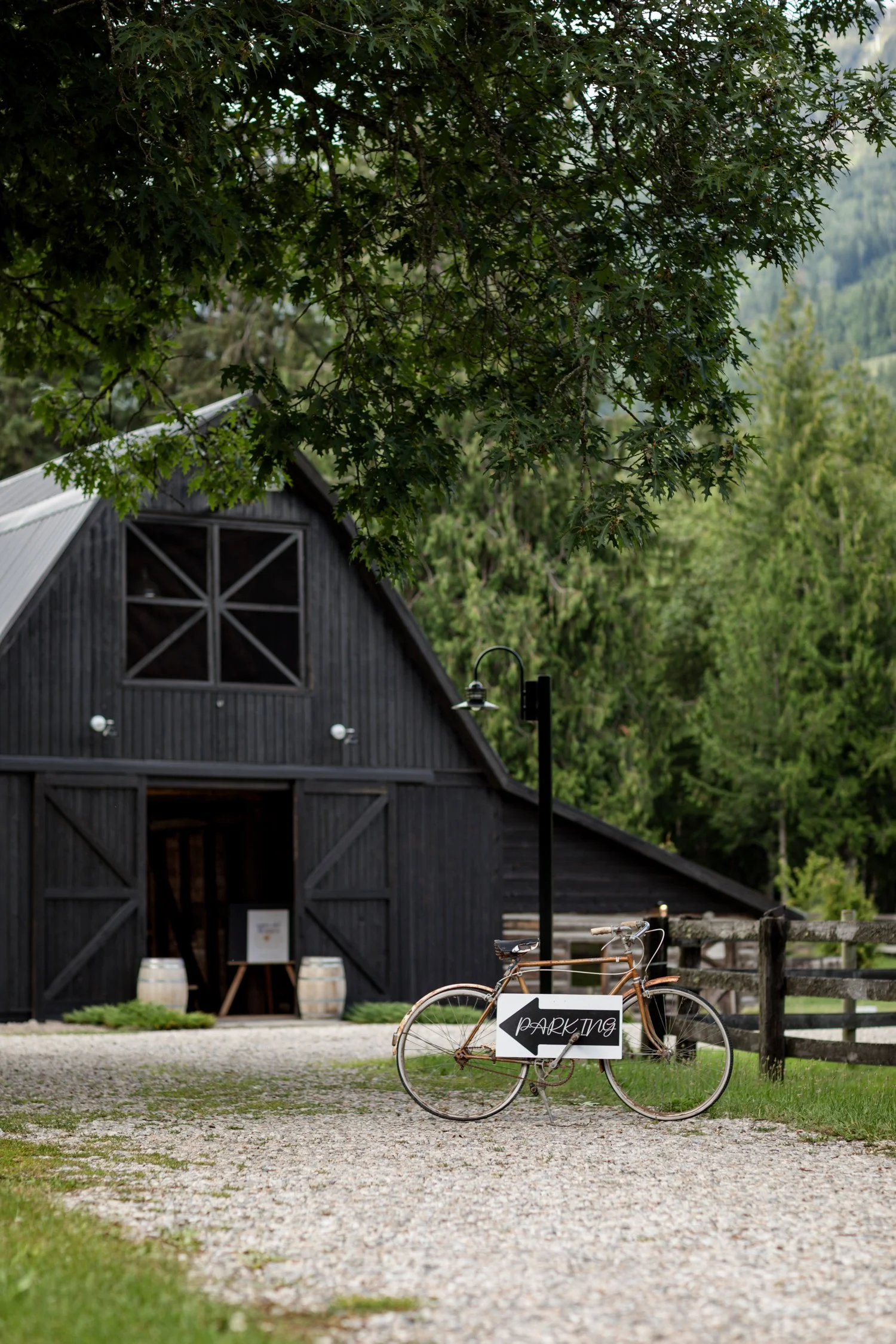 A rustic wooden barn with a metal roof and a large window, surrounded by lush green trees, with a bicycle parked in front and a parking sign on it, in a rural setting.