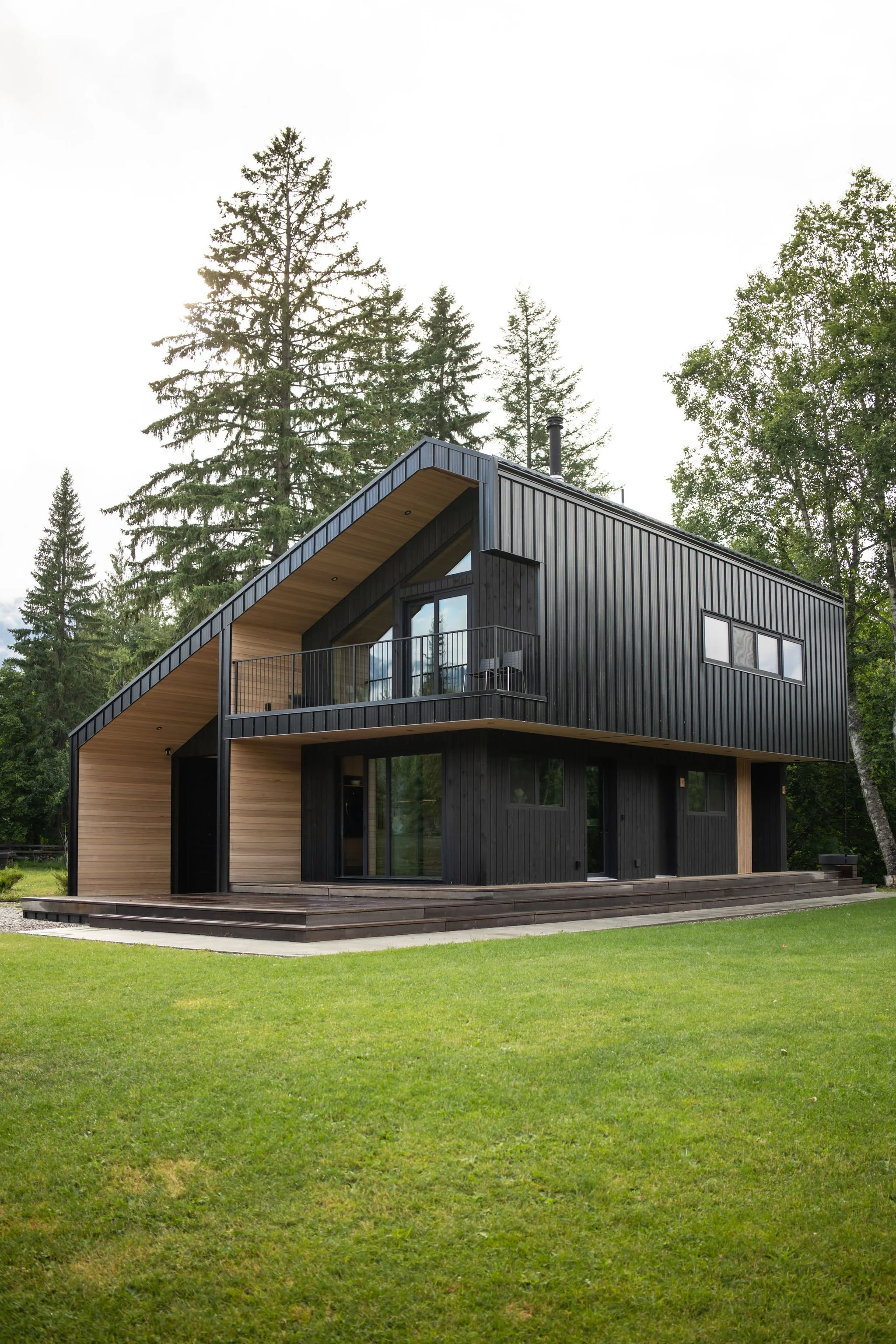 Modern black house with large windows and a wooden balcony, surrounded by green grass and tall trees in the background.