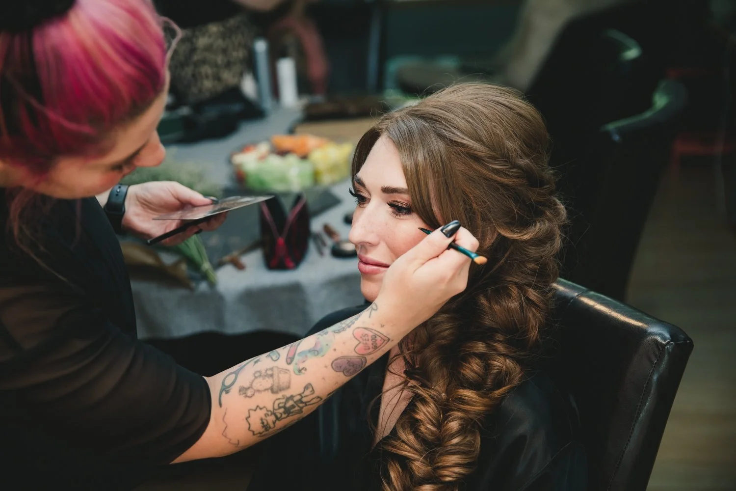A woman with brown hair is getting her makeup done by a makeup artist with pink and red hair. The makeup artist is applying eye makeup, and the woman is sitting in a black chair with her eyes closed.